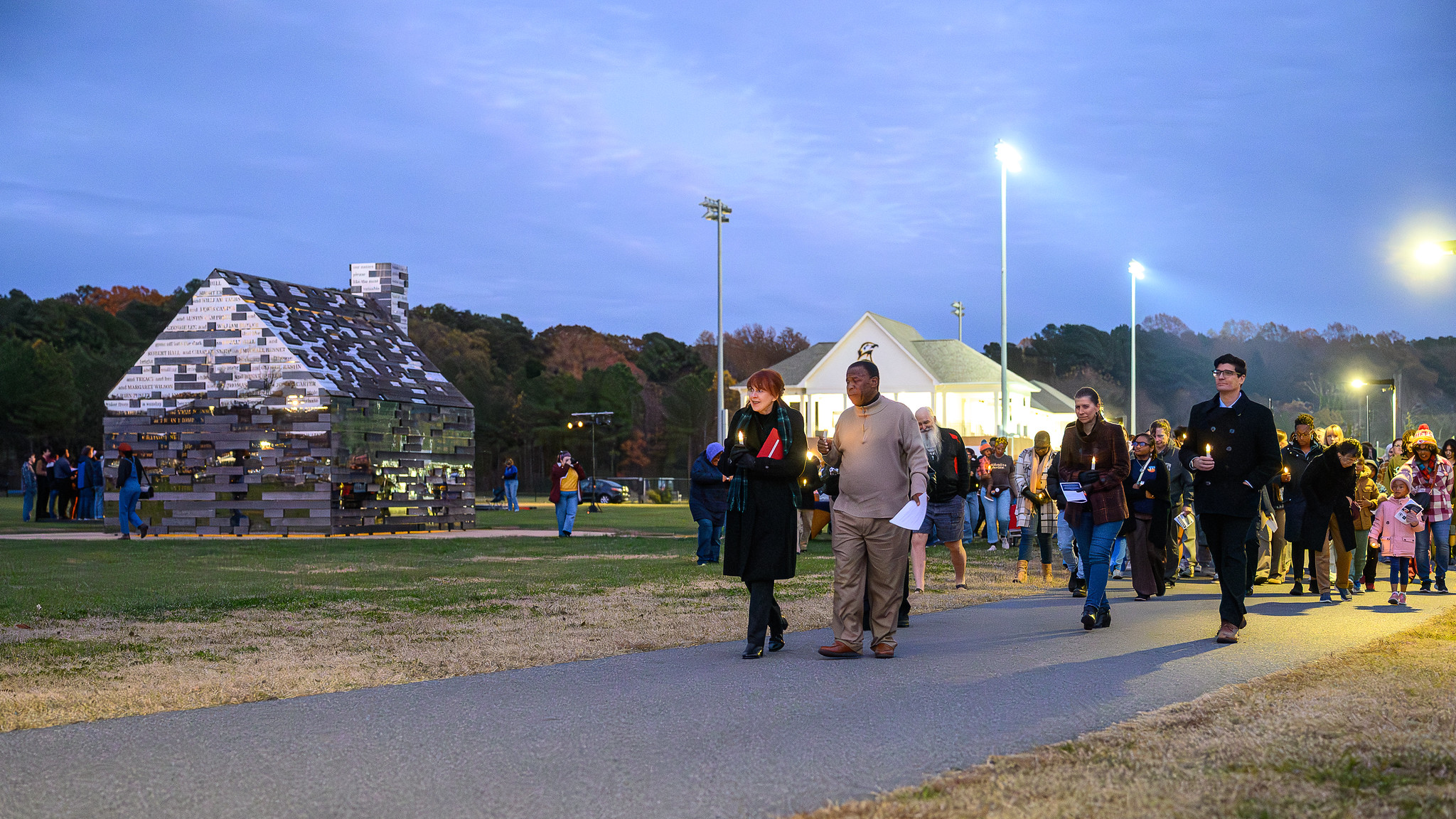 A group of people walk on a paved path in a park at dusk, with a metallic house-shaped structure and pavilion visible in the background.