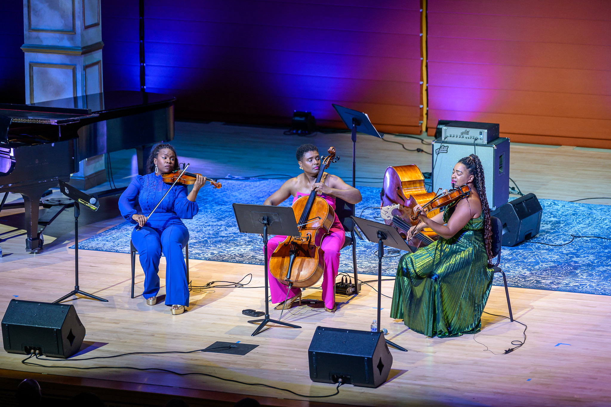 Three musicians perform on stage, playing violin, cello, and viola. They are seated with music stands on a lit wooden floor with a rug and stage lights in the background.