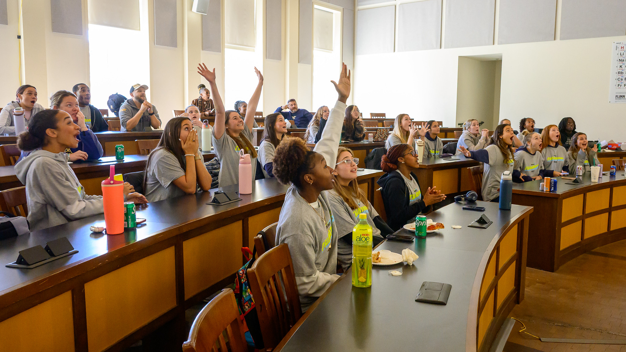 Students in a classroom sit at curved desks, some raising their hands excitedly while looking toward the front, with drinks and notebooks on the desks.
