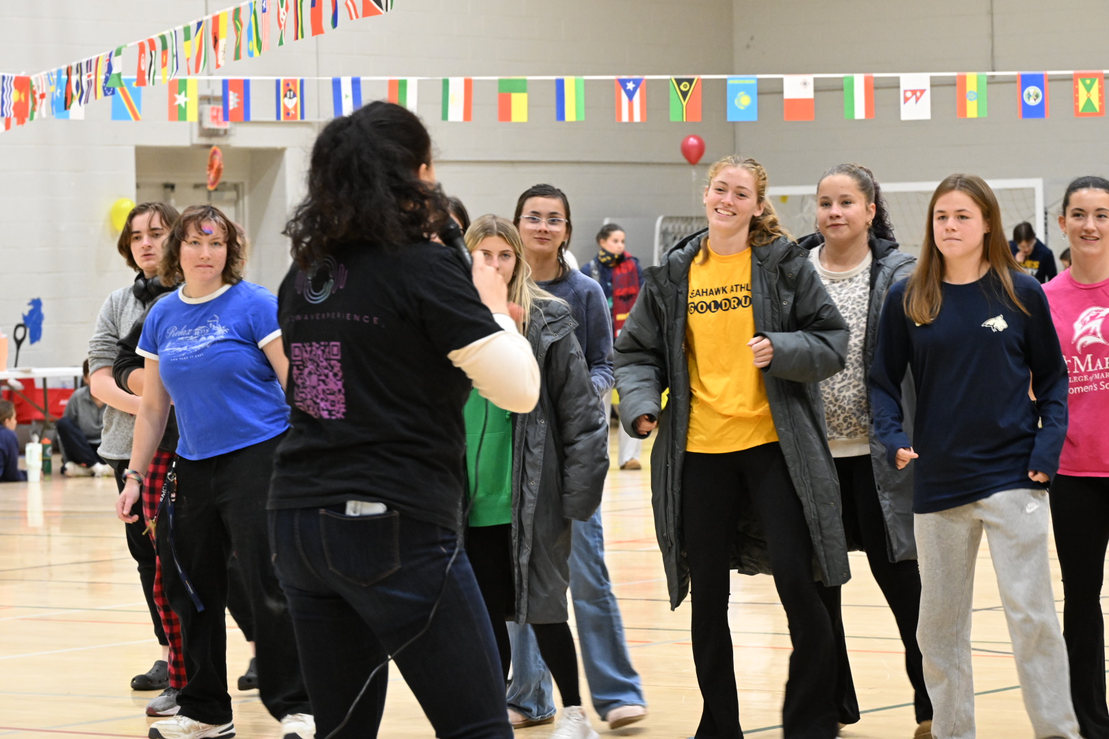 A group of young people standing in a gymnasium, listening to a woman speaking, with international flags hanging above them.