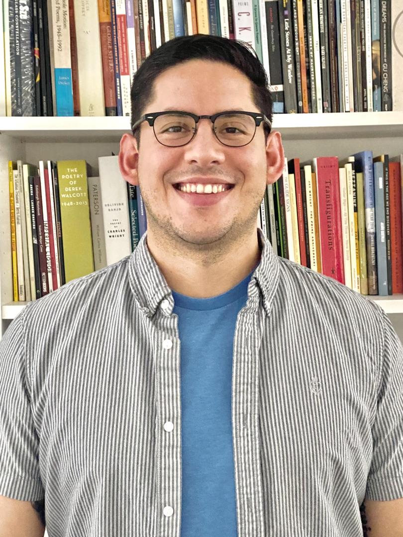 A person wearing glasses, a blue t-shirt, and a striped shirt stands smiling in front of a bookshelf filled with books.