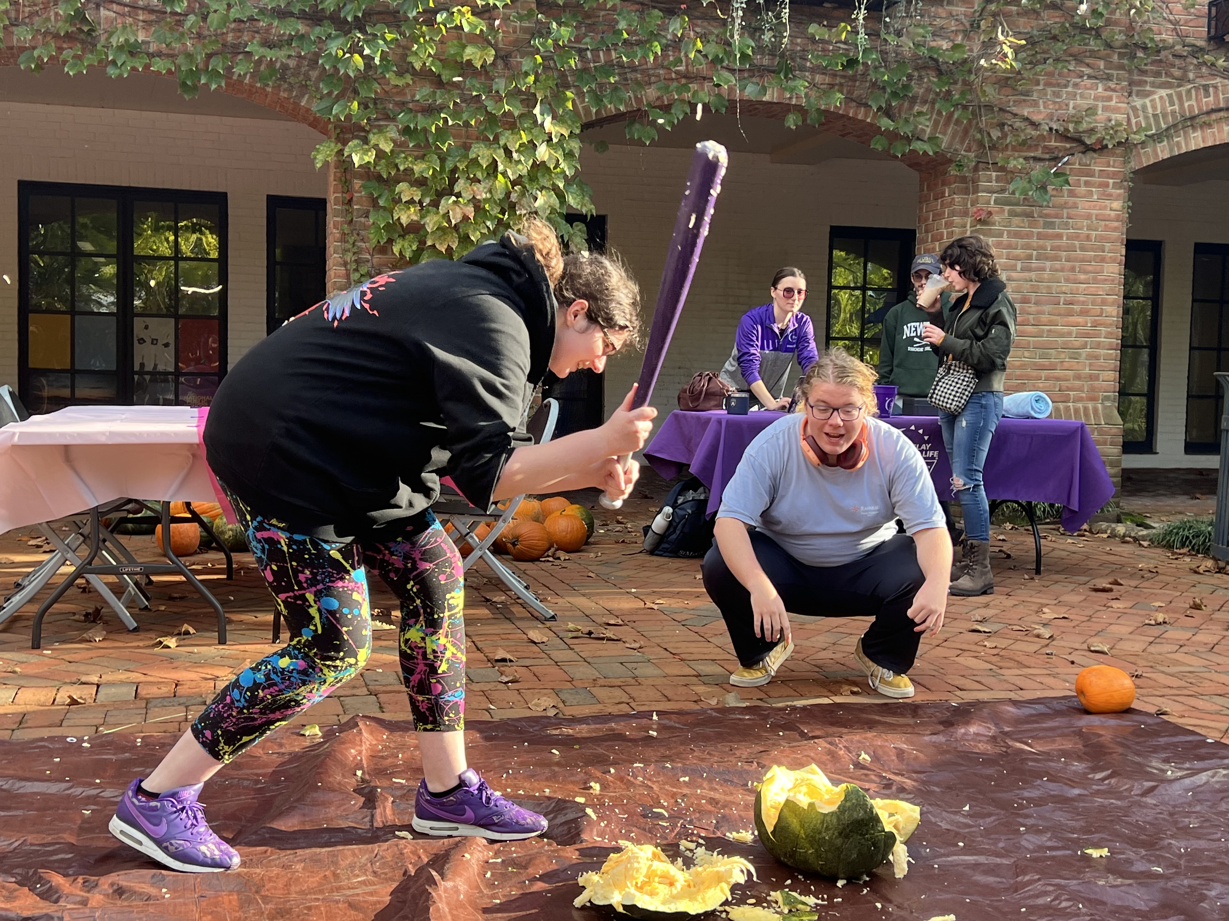A person swings a bat to smash a pumpkin on a tarp outdoors while another person crouches nearby, watching and smiling; others observe in the background.