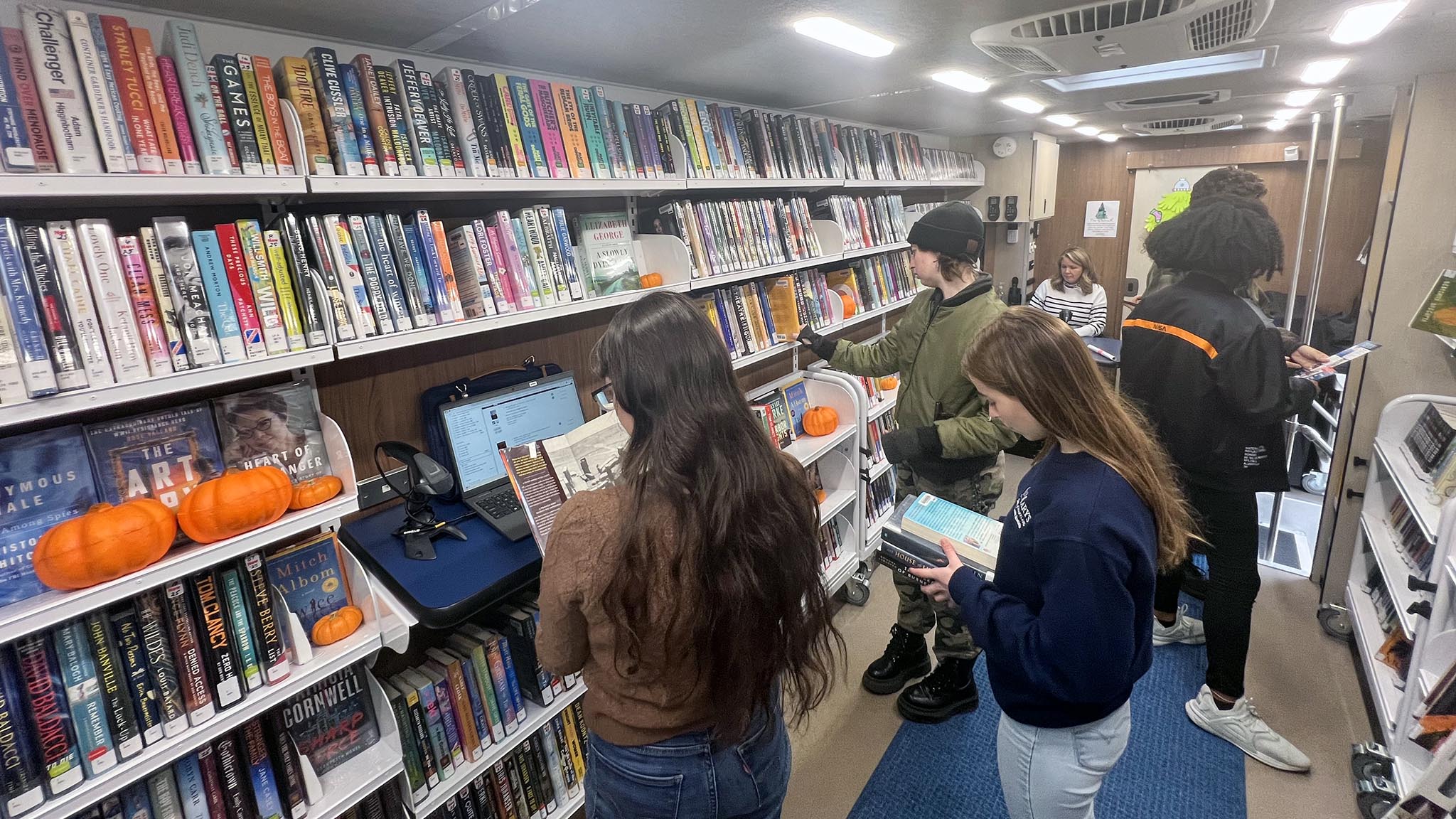 Four people browse and read books inside a mobile library filled with colorful book spines and several small pumpkins on the shelves.