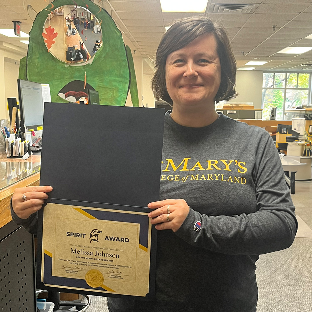 A person stands indoors holding an open certificate folder displaying a Spirit Award for Melissa Johnson. The person is smiling and wearing a St. Mary's College of Maryland shirt.