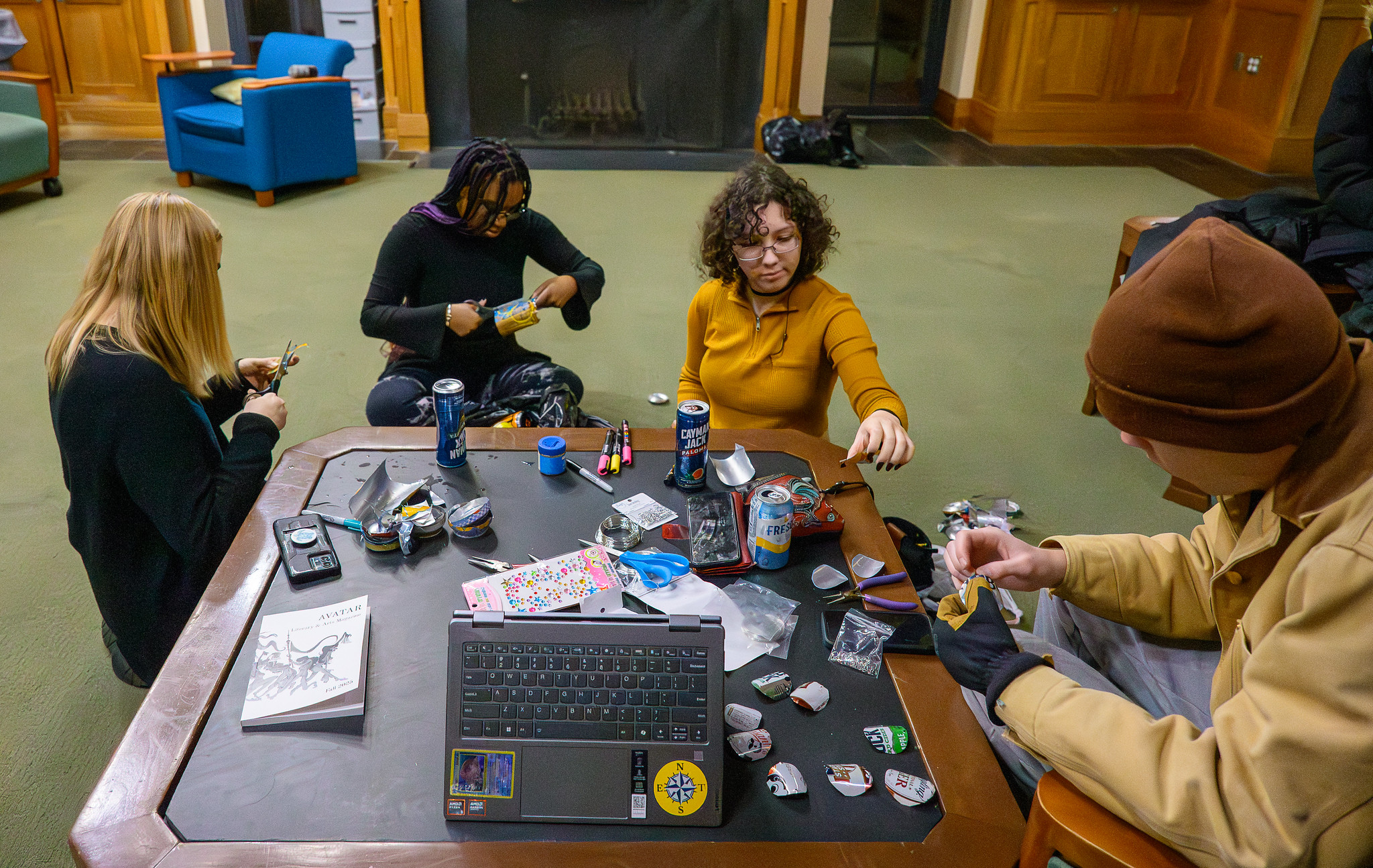 Four people sit around a table working on arts and crafts with various supplies, drinks, a laptop, and books in a lounge or common room setting.
