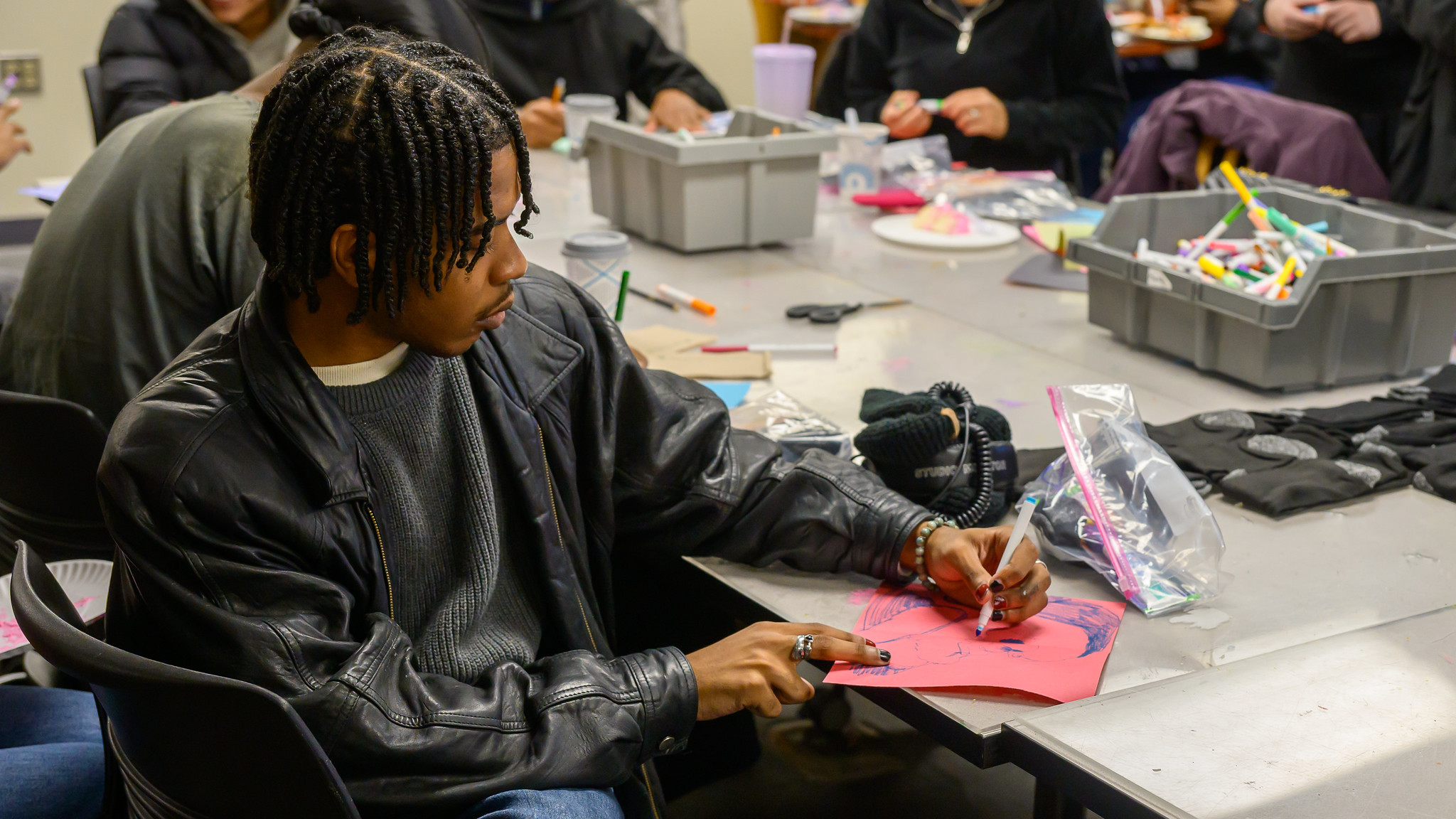 A person sits at a table drawing on red paper with markers, surrounded by art supplies and other people working on similar activities.