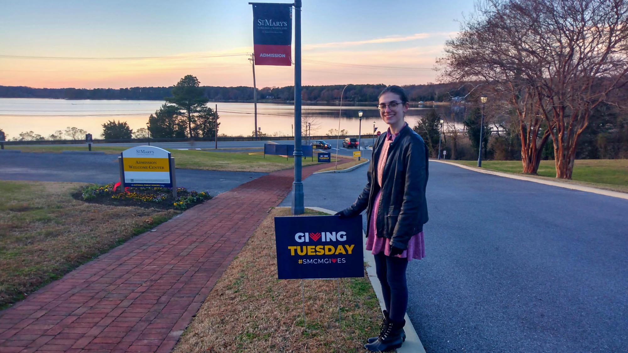 A person stands on a sidewalk holding a "Giving Tuesday #SMCMGIVES" sign near a campus entrance with a river and trees in the background at sunset.