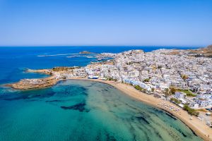 Aerial view of a coastal town on Naxos with white buildings, a sandy beach, and clear blue-green water under a clear sky.