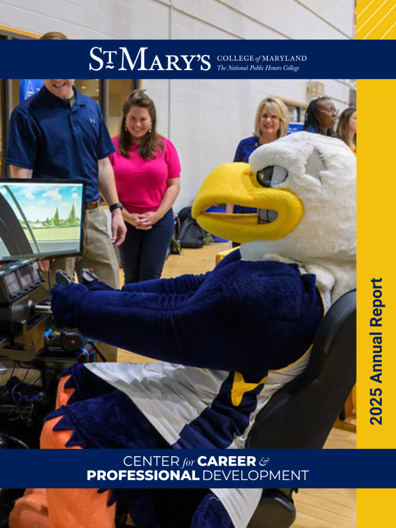 A person in an eagle mascot costume sits at a driving simulator while several people stand nearby, smiling, in a gymnasium setting.