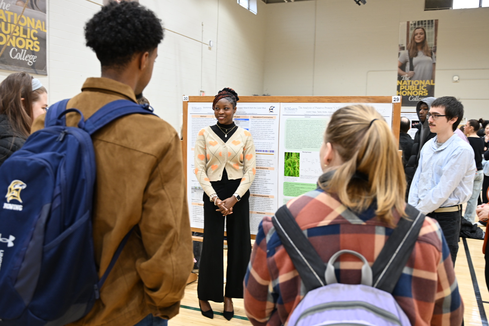 A student stands in front of a science poster presentation, speaking to a small group of people in a gymnasium setting.