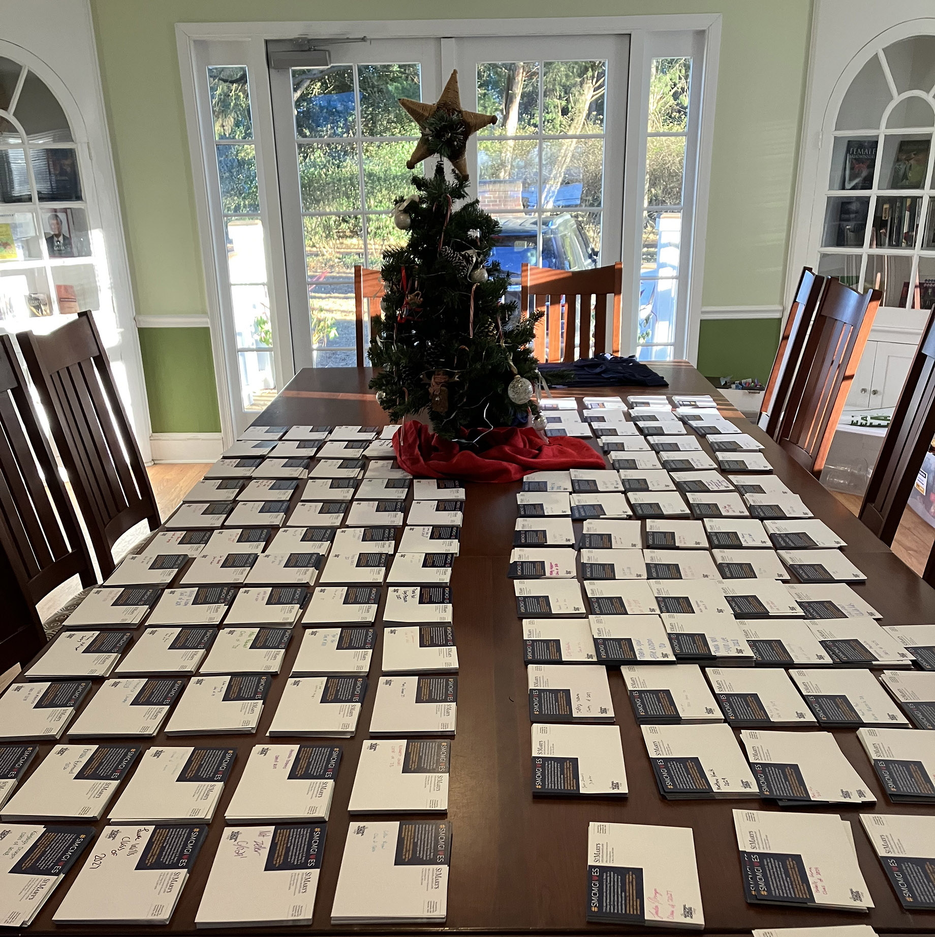 A dining table covered with rows of cards or envelopes, with a decorated Christmas tree centerpiece and sunlight streaming in through glass doors.