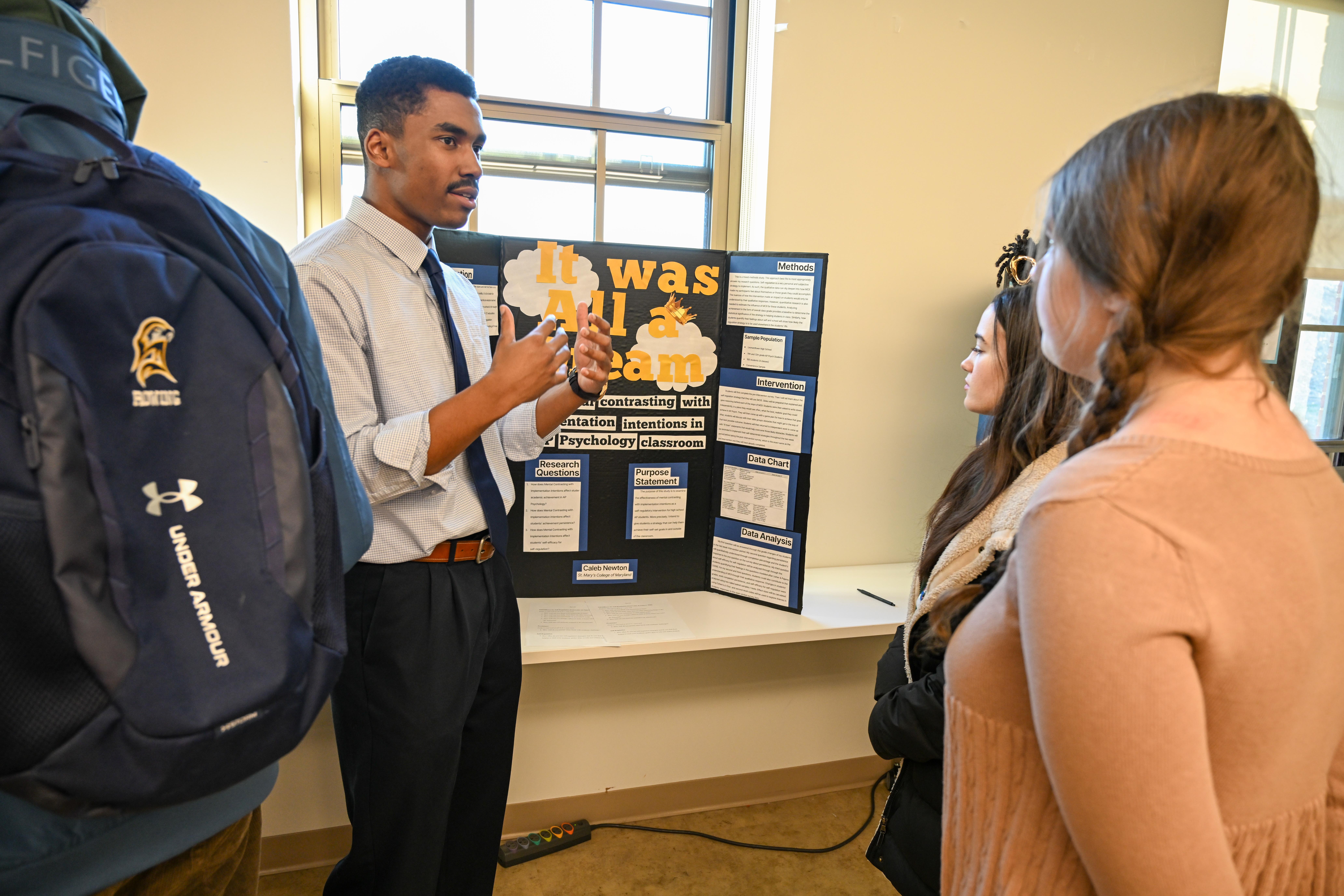 A man presents a project titled "It was All a Dream" on a trifold board to three people in a classroom setting.