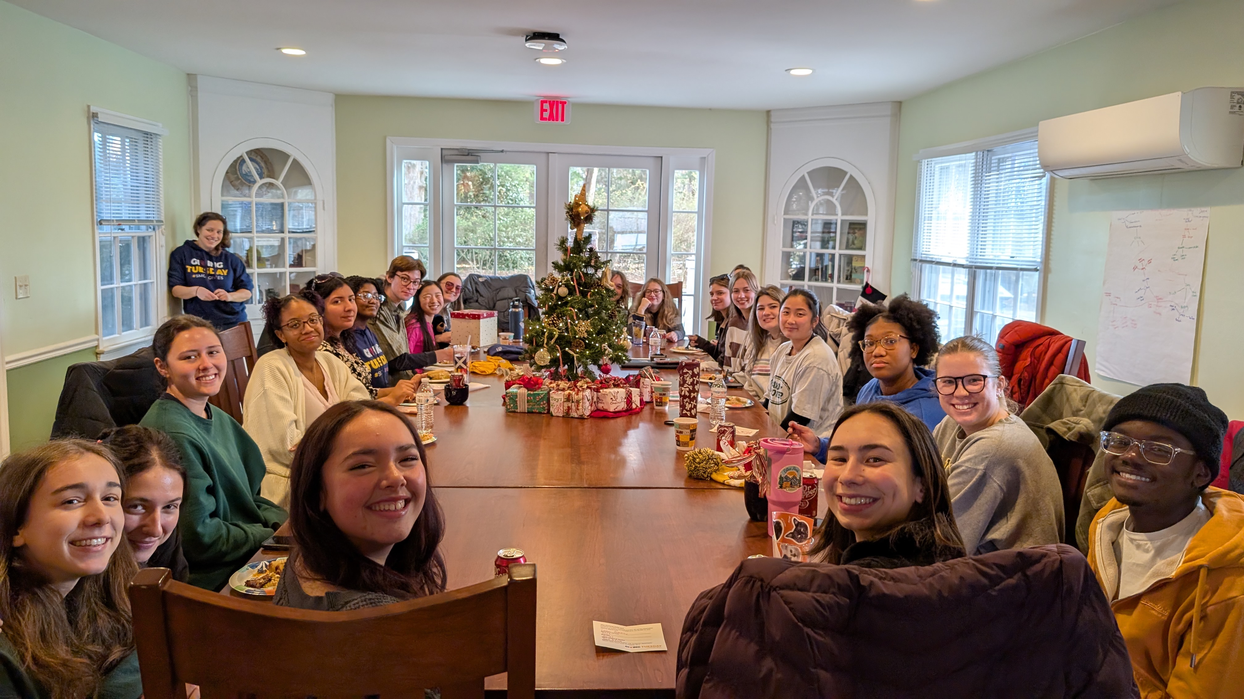 A group of people sit around a large table with food, drinks, and a small decorated Christmas tree in the center, gathered in a bright, cozy room.