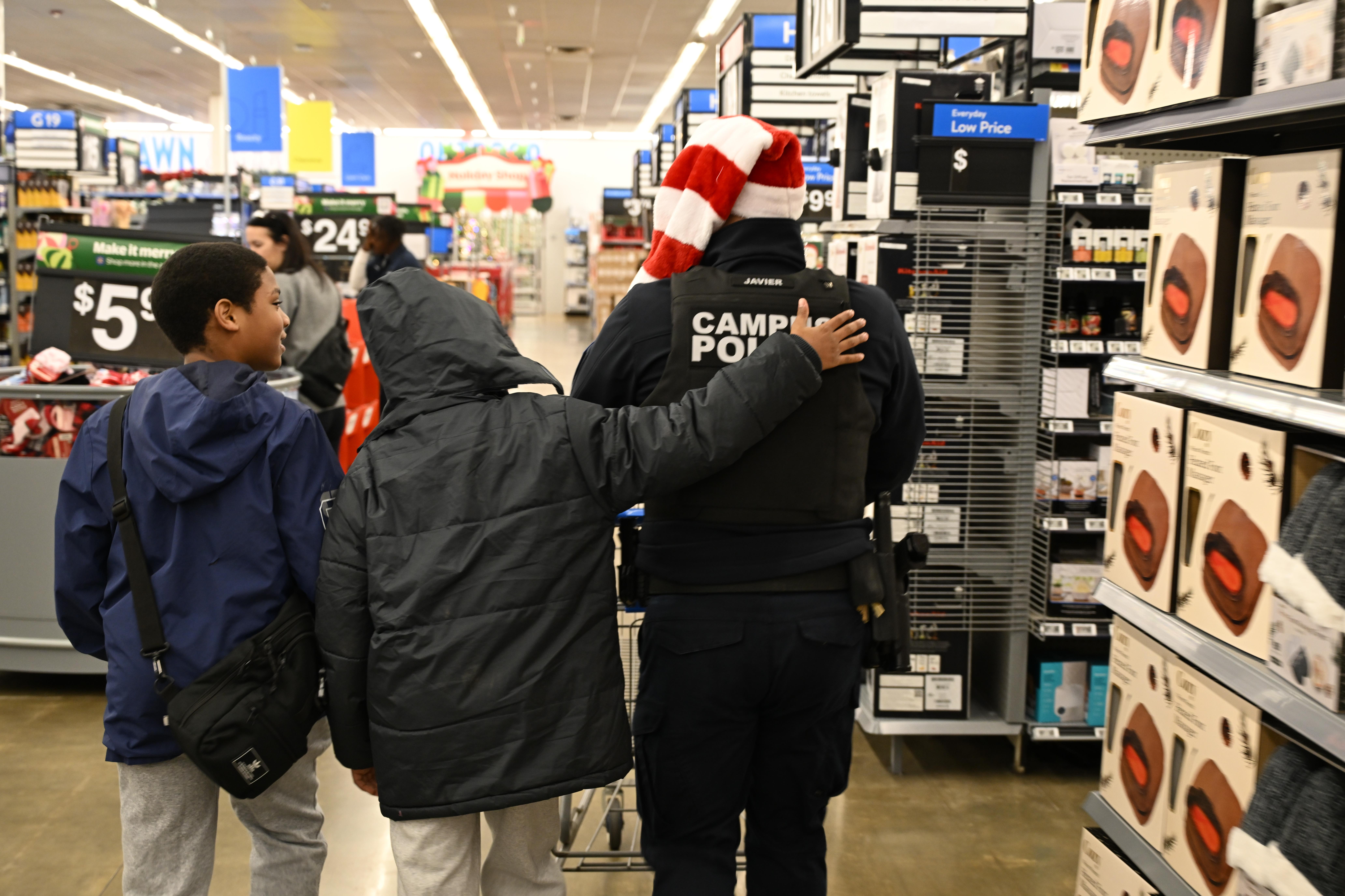 Two boys walk with a campus police officer wearing a Santa hat in a store aisle lined with boxed products.