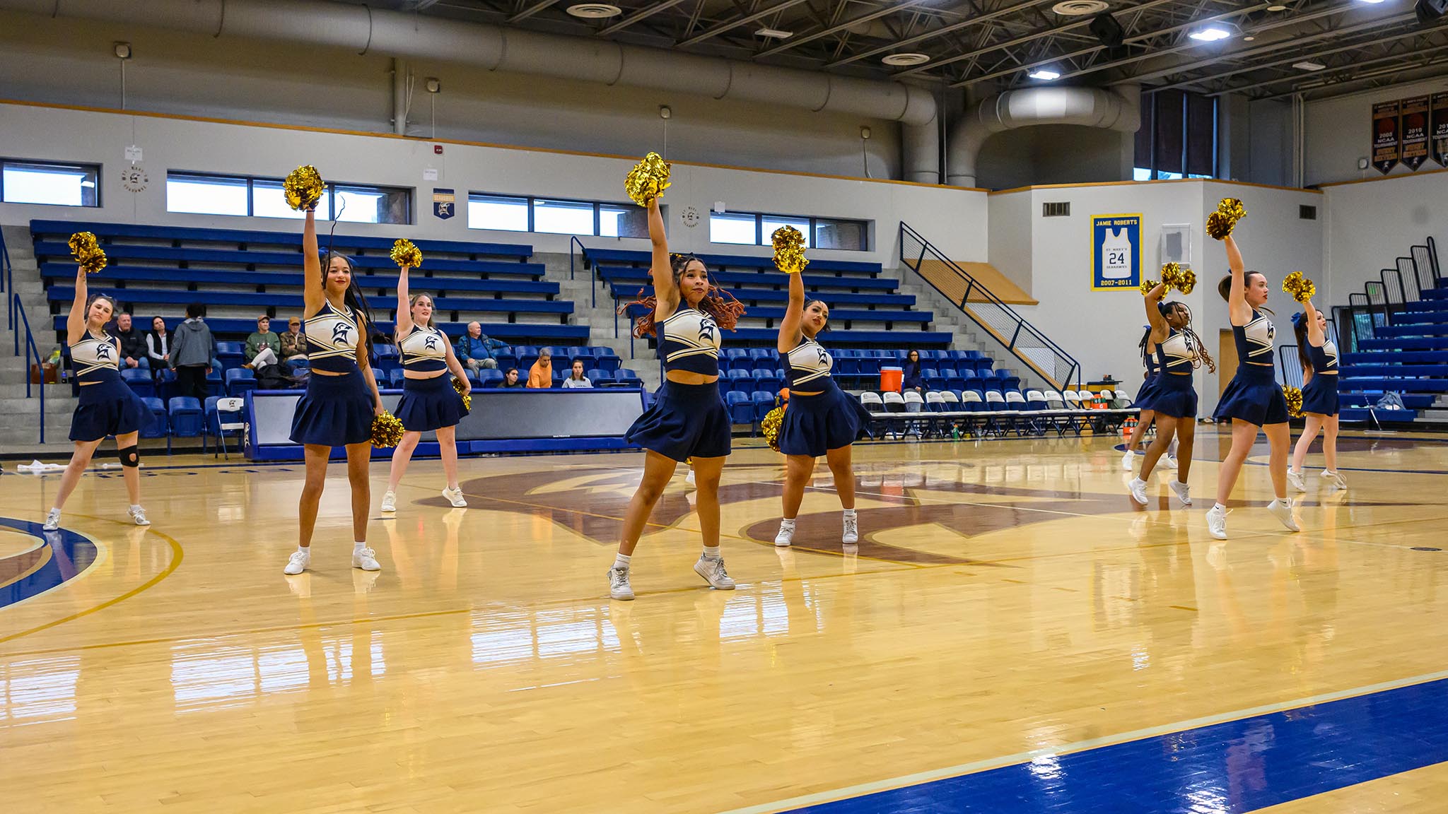 A group of cheerleaders in uniform perform a routine with gold pom-poms on a basketball court in a gymnasium.