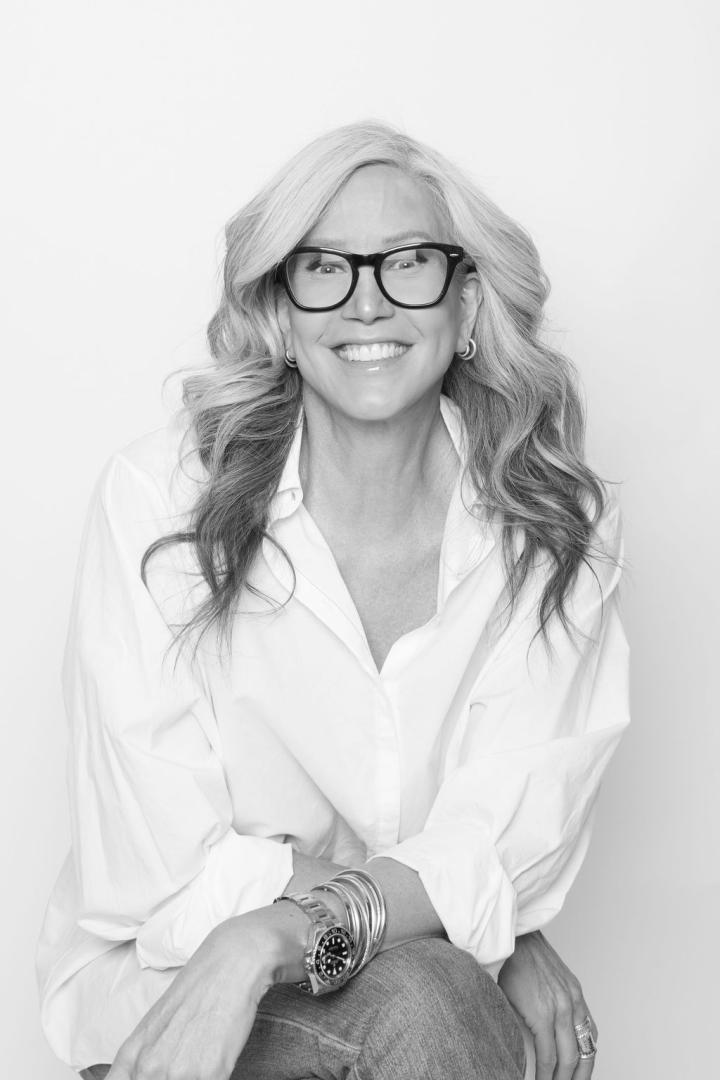 A woman with long wavy hair, wearing glasses, a white shirt, and bracelets, sits smiling with her hands resting on her knee in a black and white photo.