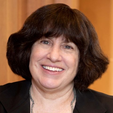 A woman with short, dark brown hair and a black blazer smiles at the camera against a blurred, warm-toned background.