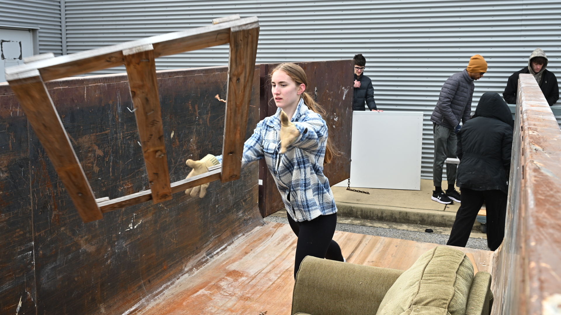 A woman throws a wooden chair into a large dumpster while several other people clear out items nearby.
