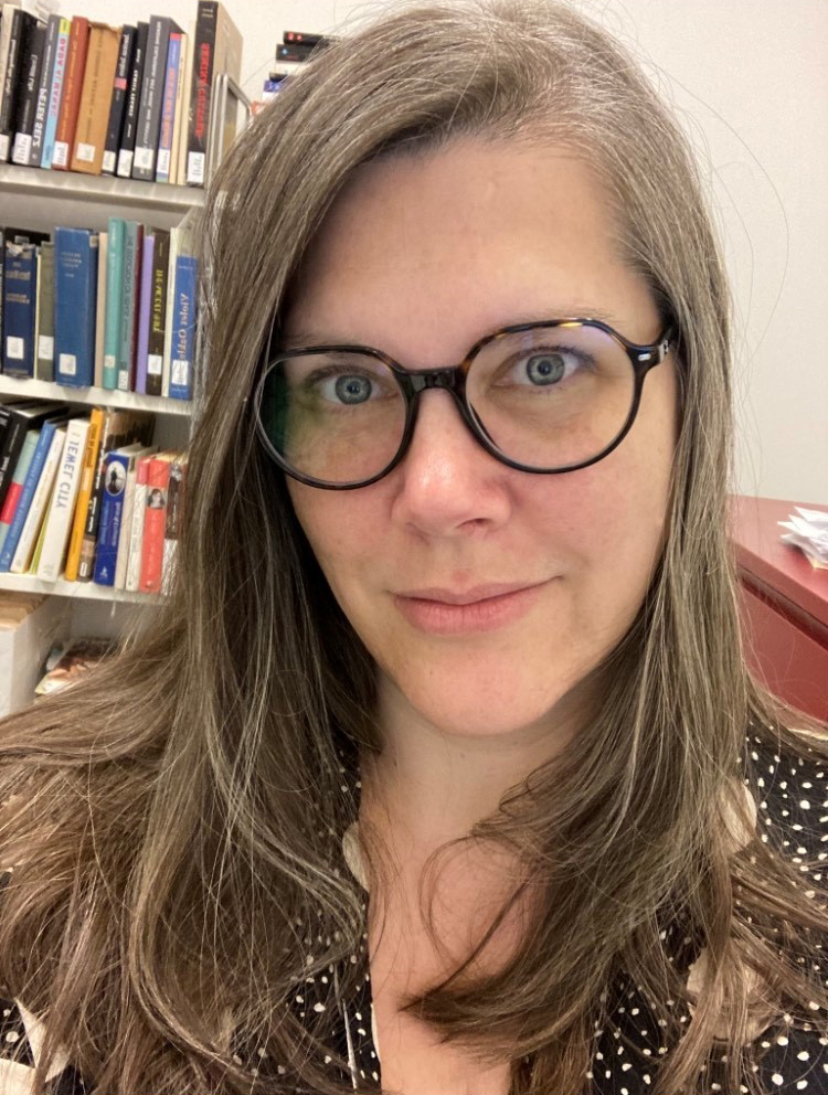 A woman with long brown hair and glasses looks at the camera, standing in front of a bookshelf filled with books in an office setting.