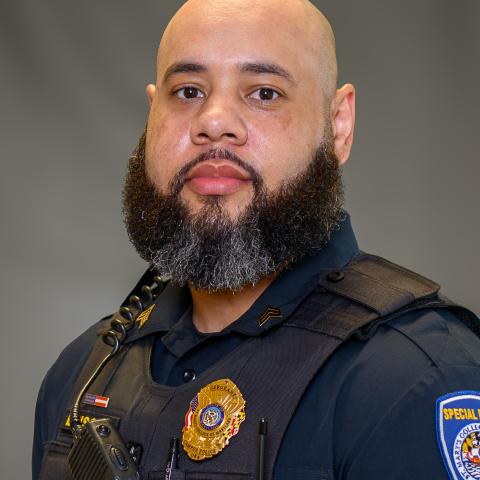 A police officer with a beard wearing a uniform, badge, body armor, and radio, posing against a plain background.