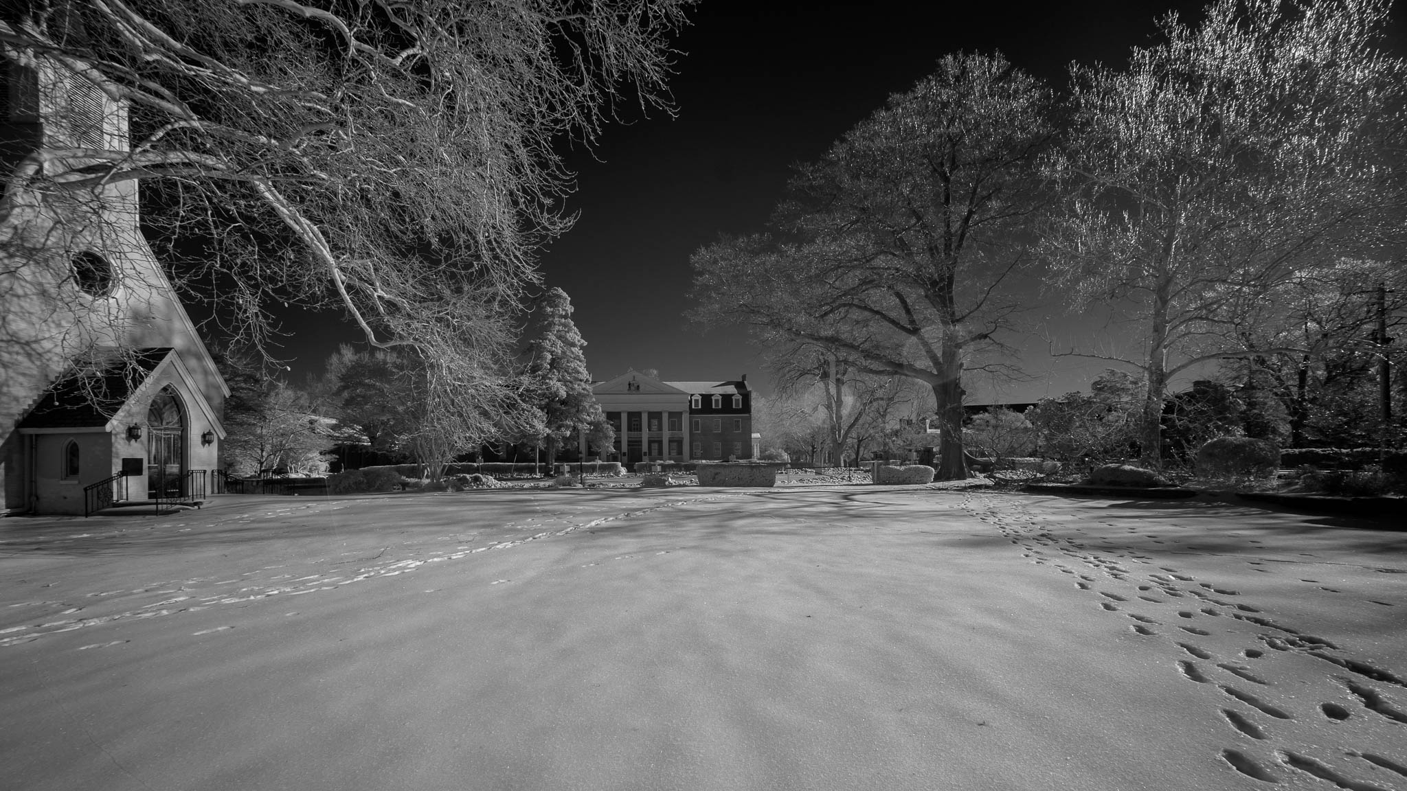A snow-covered landscape with footprints leading to and from a church building on the left and a large house in the background, surrounded by trees under a clear sky.