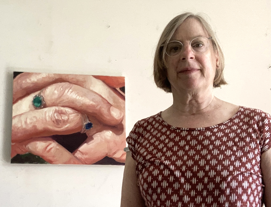 A person wearing glasses and a patterned shirt stands indoors next to a painting of two hands with rings on the fingers.