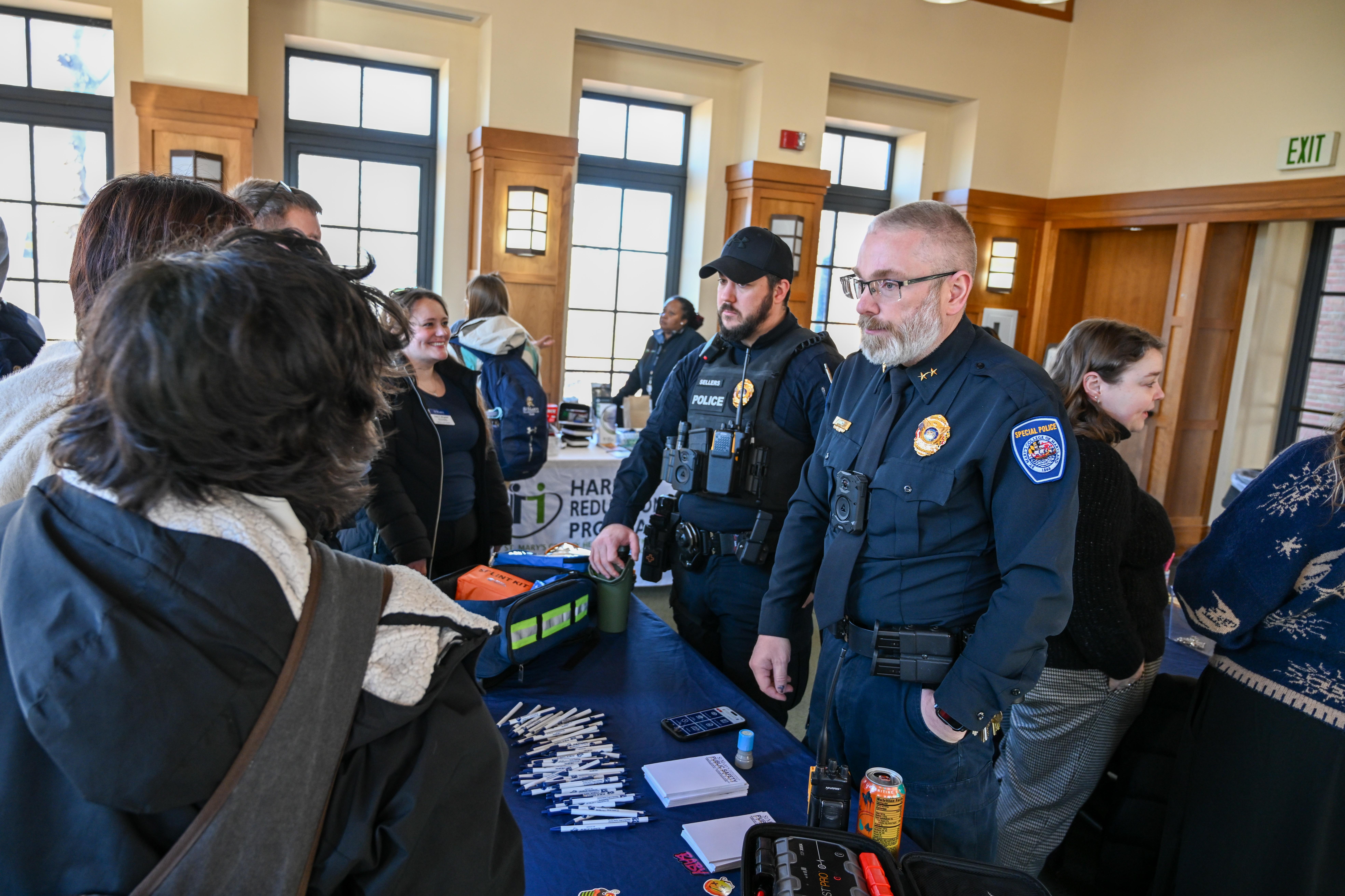 Two uniformed police officers speak to a group of people at an indoor event; informational materials and pens are displayed on the table in front of them.