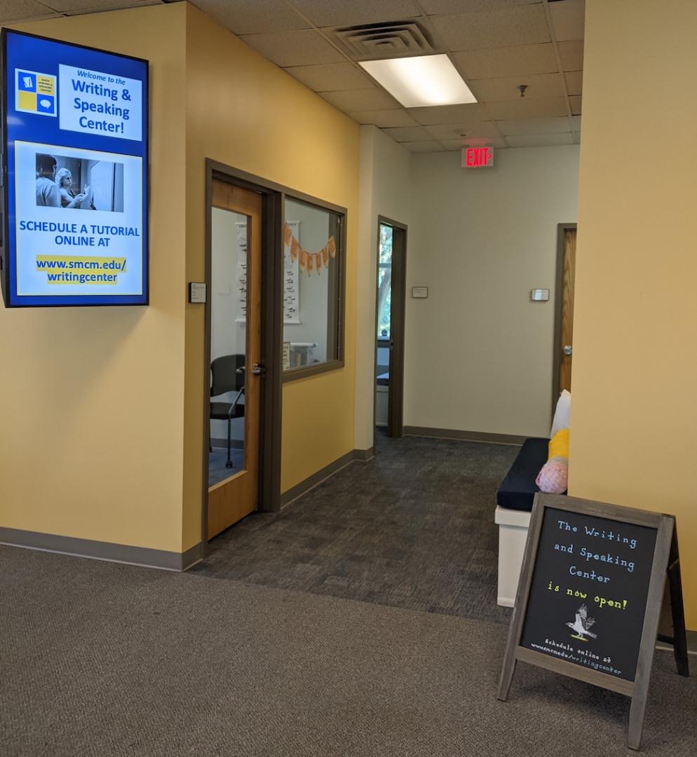 Hallway with office and tutor room doors on the left, a digital sign for the Writing & Speaking Center, and a chalkboard sign stating the center is now open.