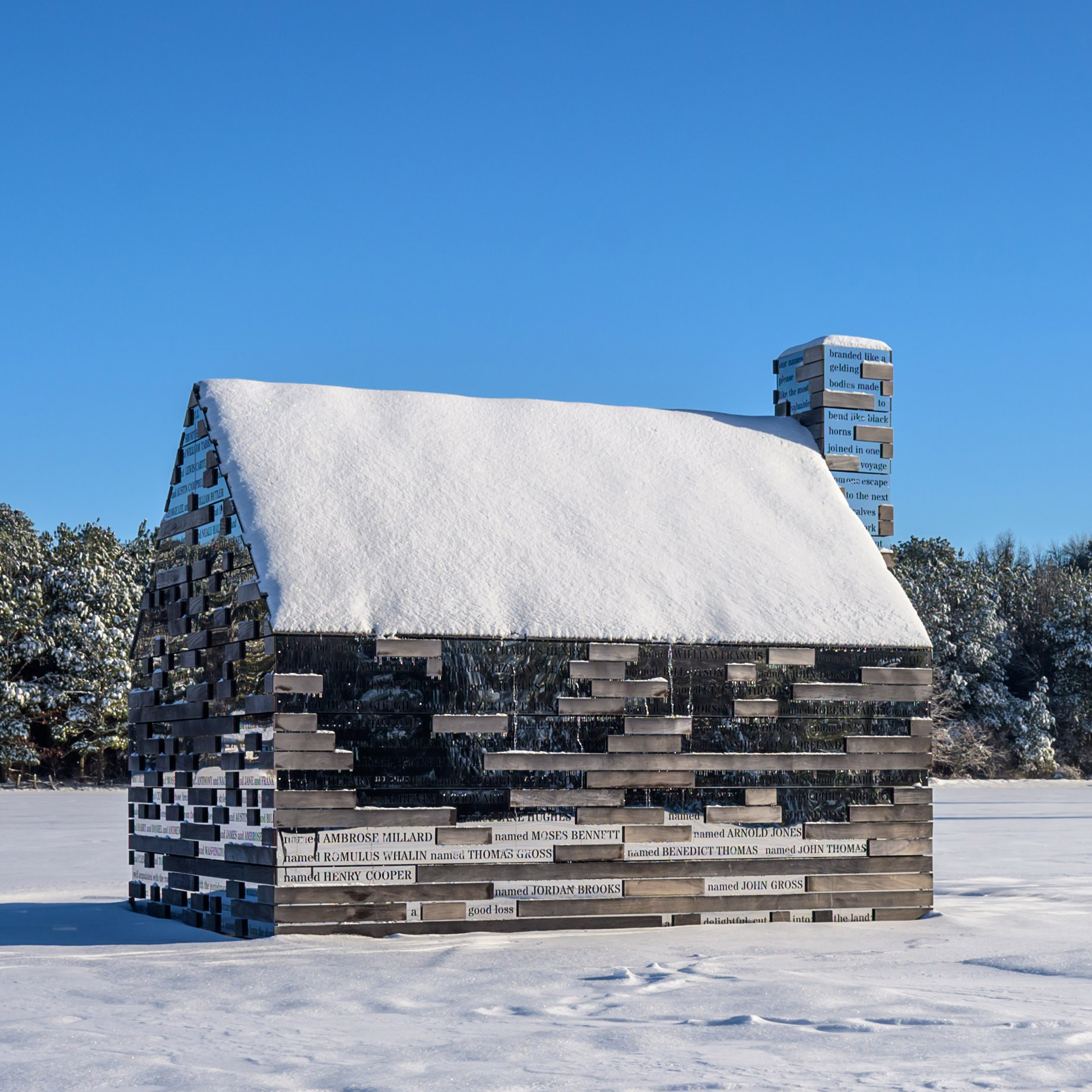 A small wooden cabin covered in snow, with names engraved on its exterior planks, stands in a snowy landscape under a clear blue sky.