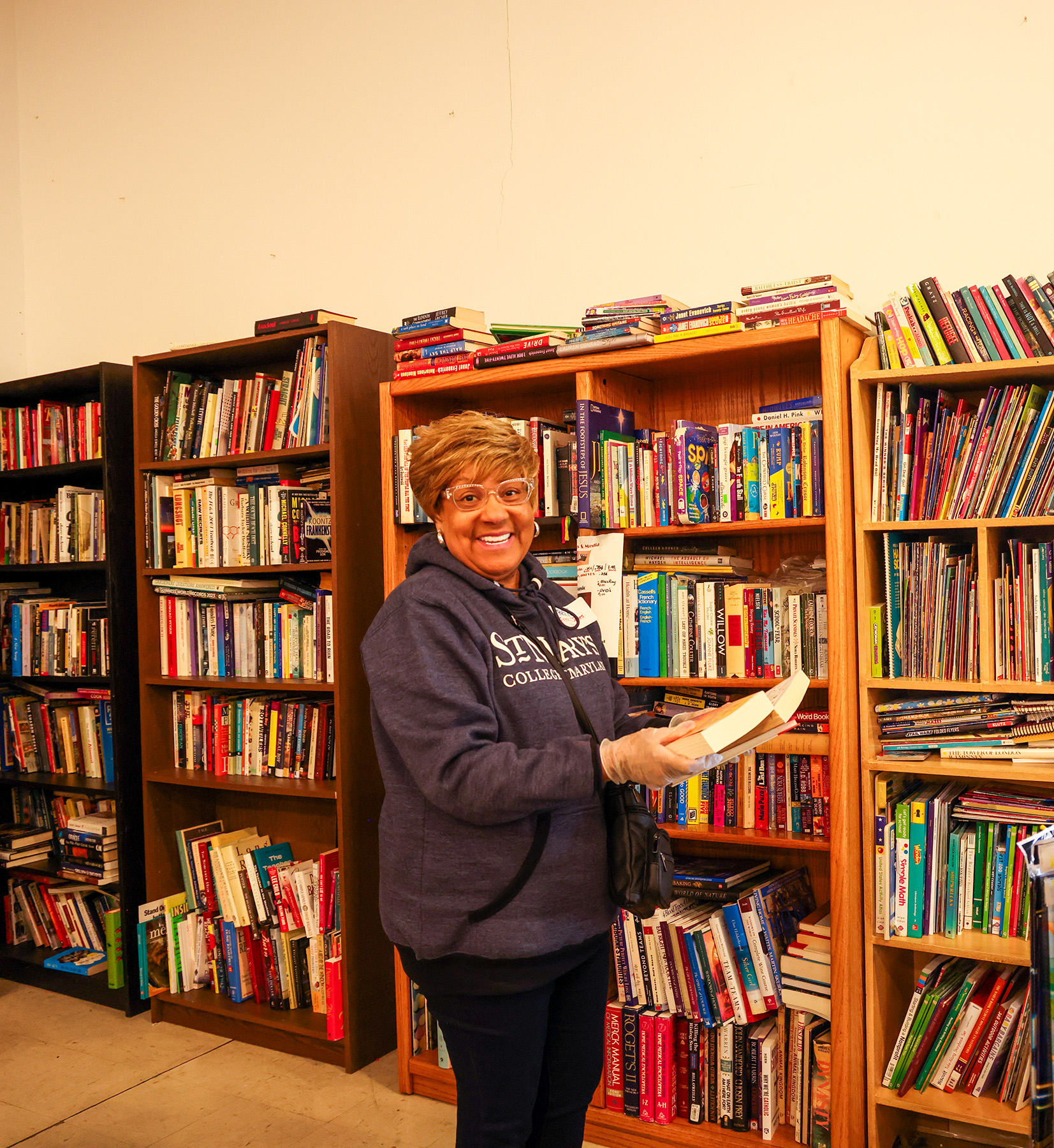 A woman wearing glasses and a hoodie stands in front of bookshelves, holding a book and smiling at the camera in a library or bookstore.