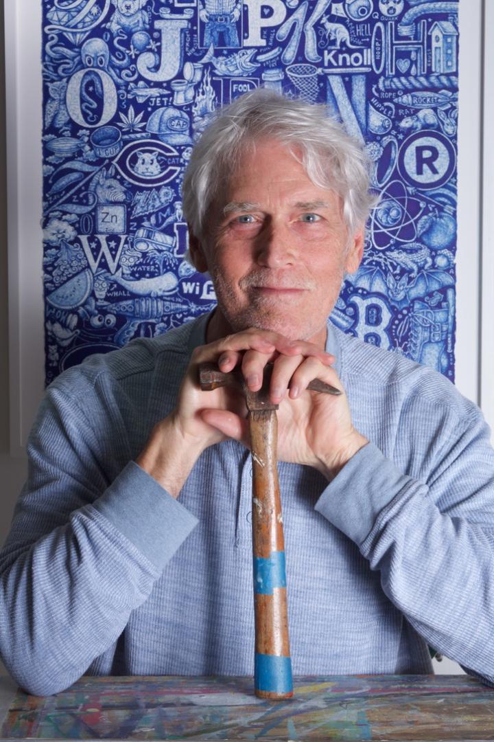 An older man with gray hair sits in front of a blue patterned artwork, resting his hands and chin on the handle of a worn hammer.