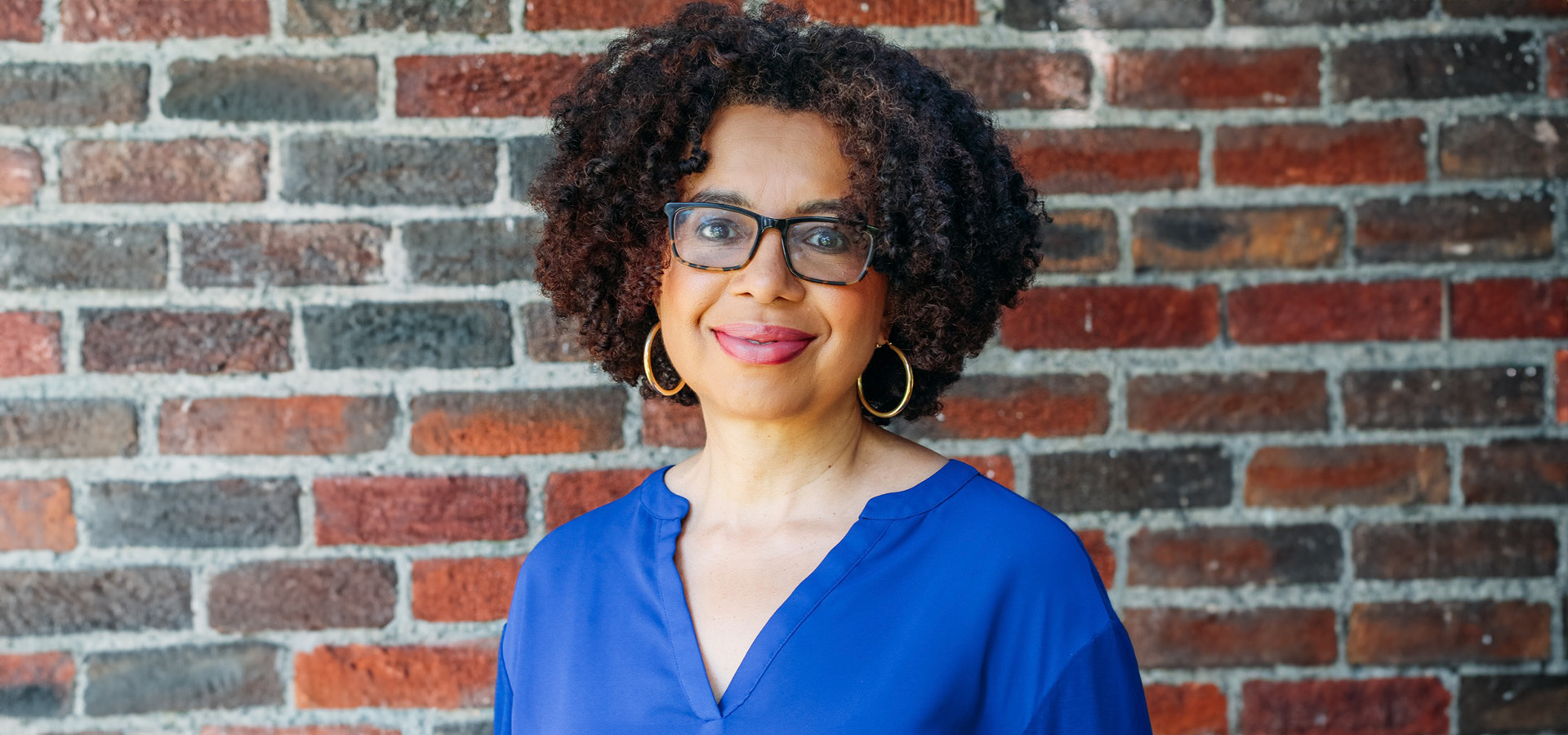 Woman with curly hair and glasses stands in front of a brick wall, wearing a blue blouse and gold hoop earrings, smiling at the camera.