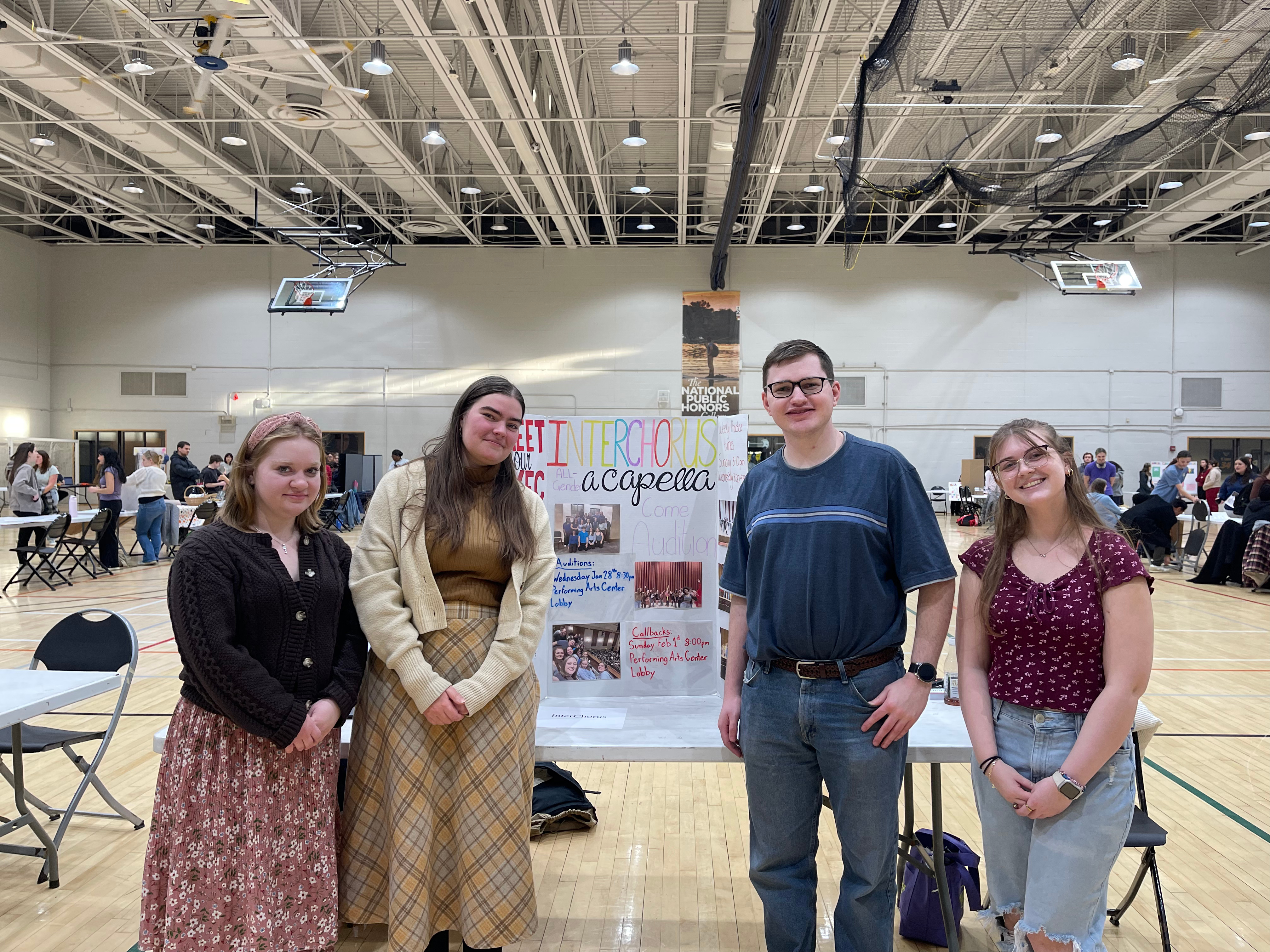 Four people stand in front of a display board labeled "Interrobang A Cappella" in a gymnasium during an indoor event. Tables and participants are visible in the background.