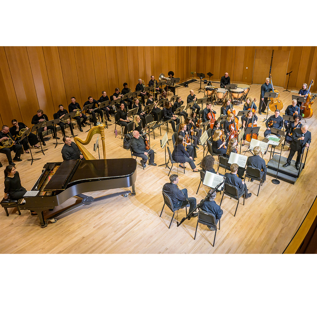 An orchestra is seated on a wooden stage with a grand piano, harp, strings, woodwinds, brass, and percussion instruments, all facing a conductor.