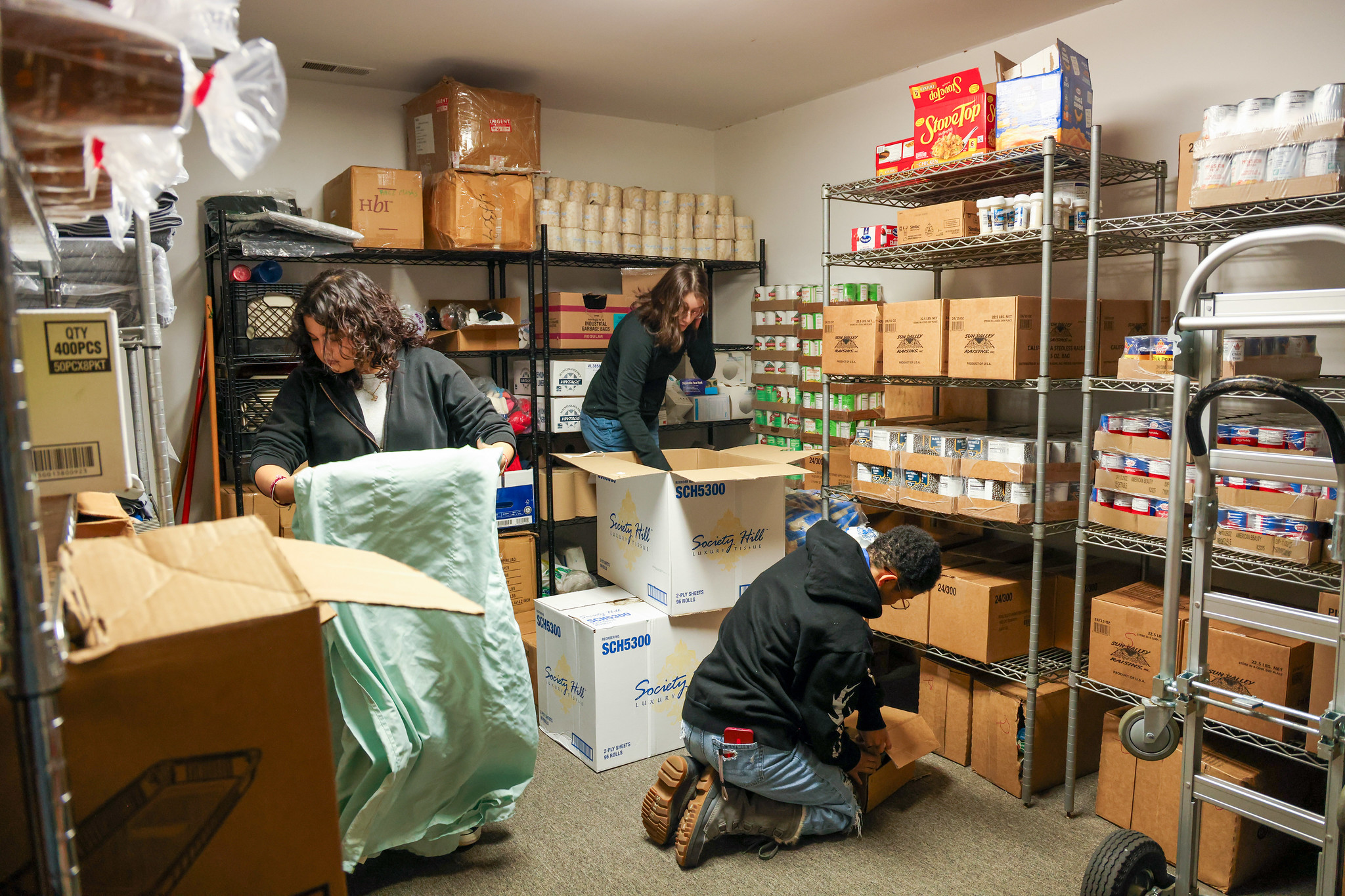 Three people organize and pack boxes of food in a storage room filled with shelves stocked with canned goods and other supplies.