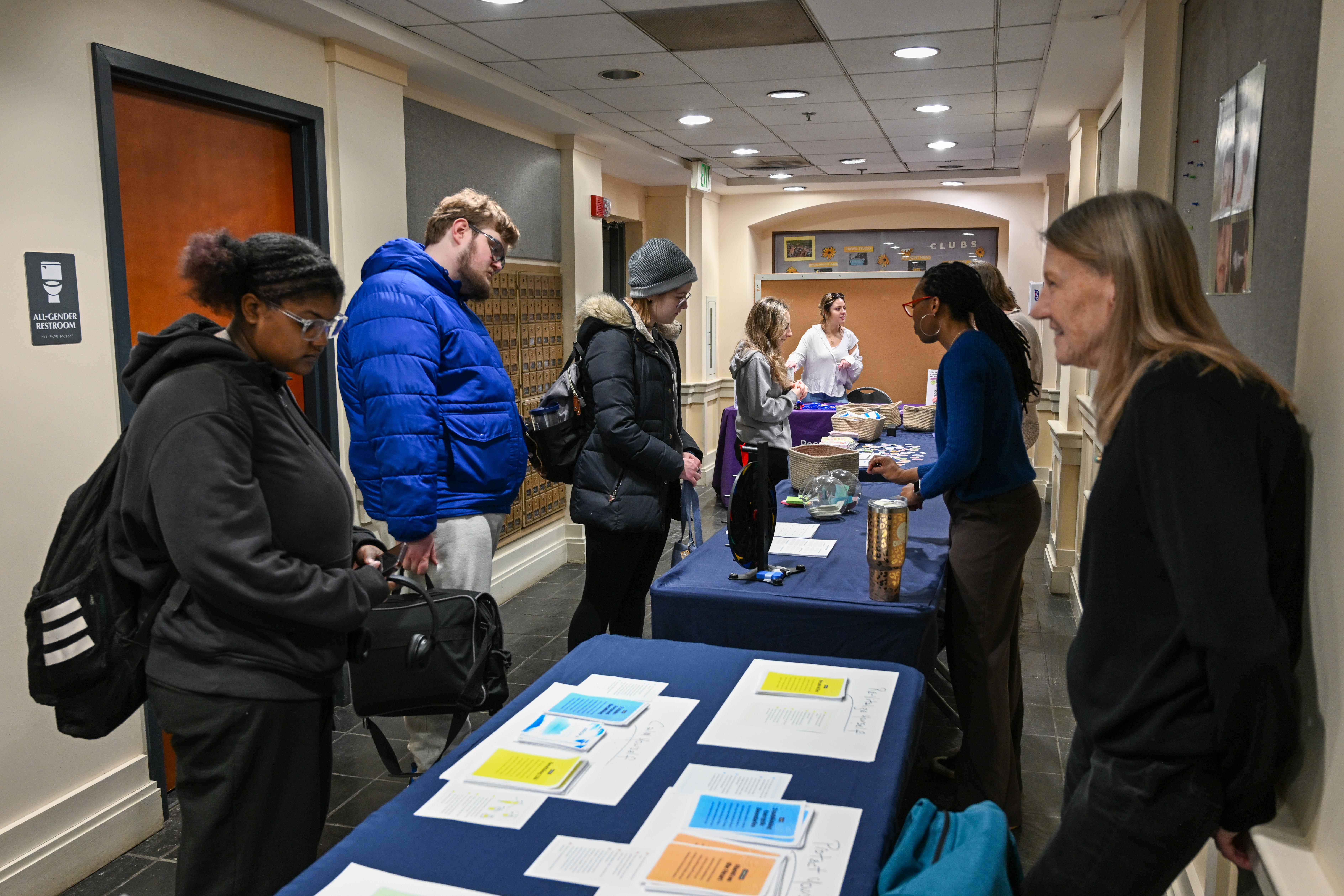 People stand at tables in a hallway, interacting with staff and picking up papers and food items; informational flyers are laid out in the foreground.