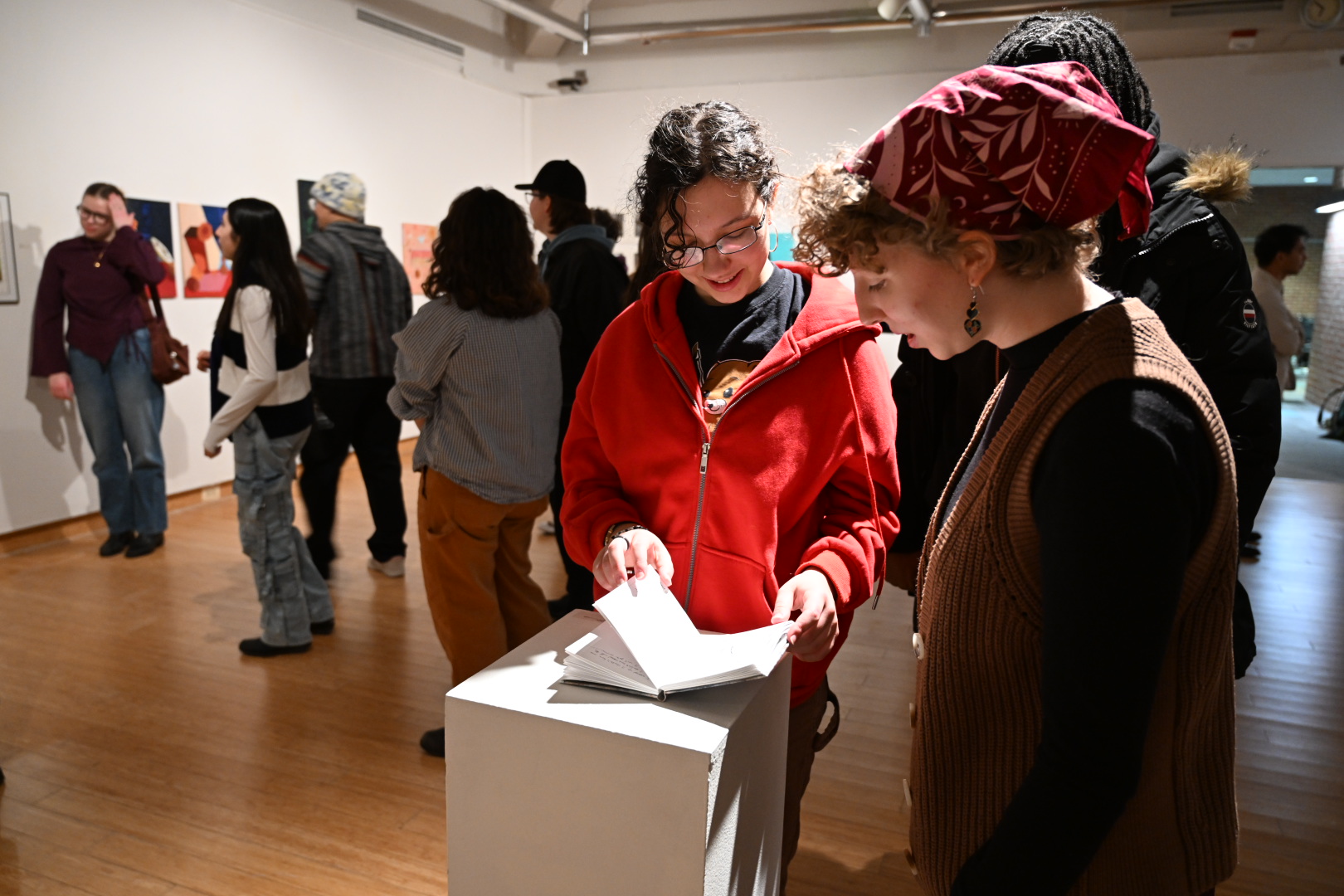 People view art and read a booklet at an indoor gallery exhibition. The room has wooden floors, white walls, and several artworks on display.