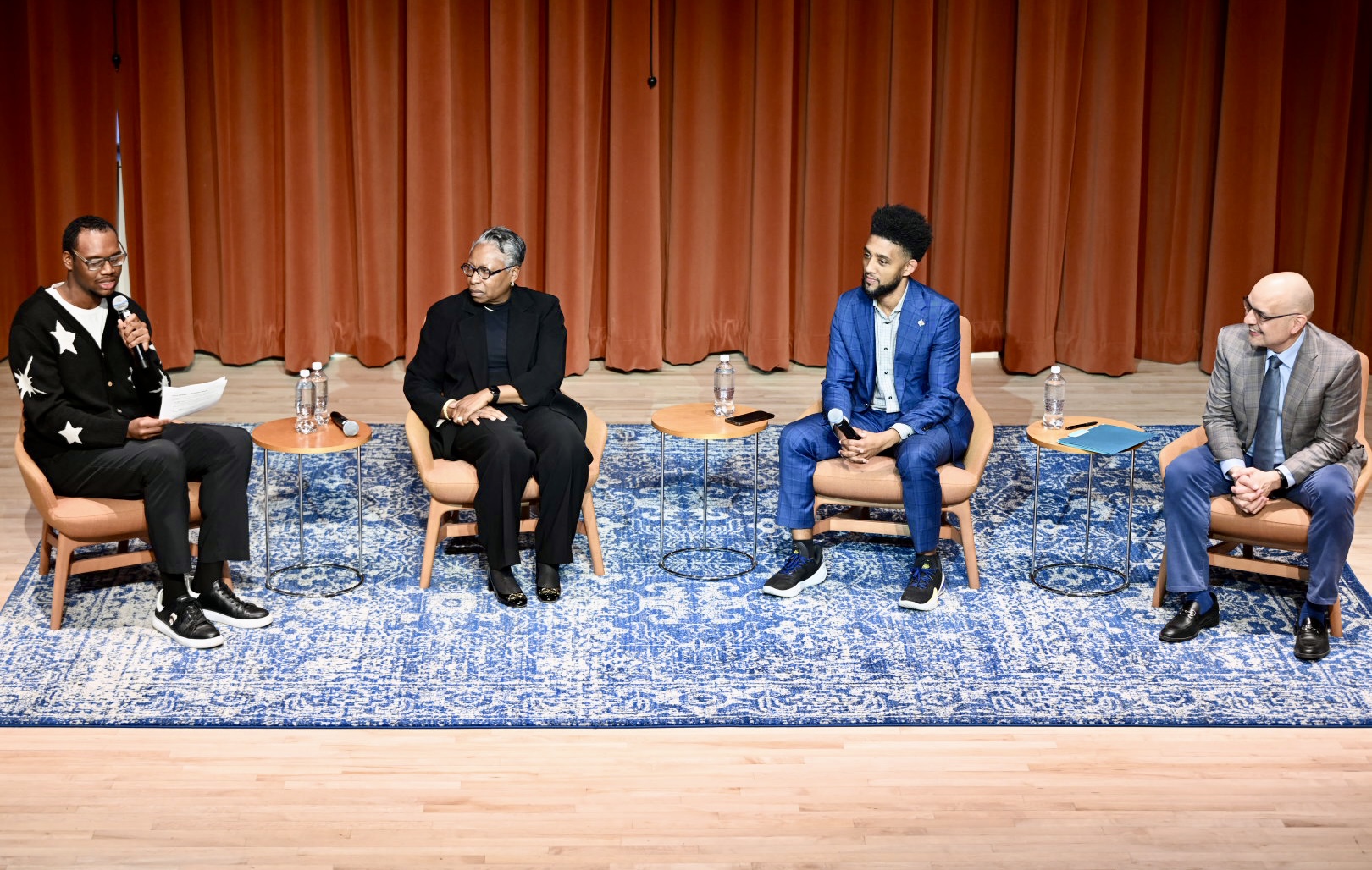 Four people sit on stage in armchairs, engaged in a panel discussion. One person is speaking into a microphone while the others listen. There are small tables with water bottles beside each chair.