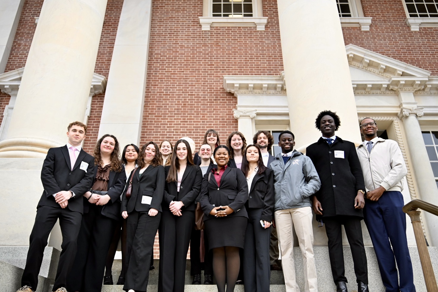 A group of thirteen people in business attire stand together on the steps of a brick building with columns, posing for a group photo.