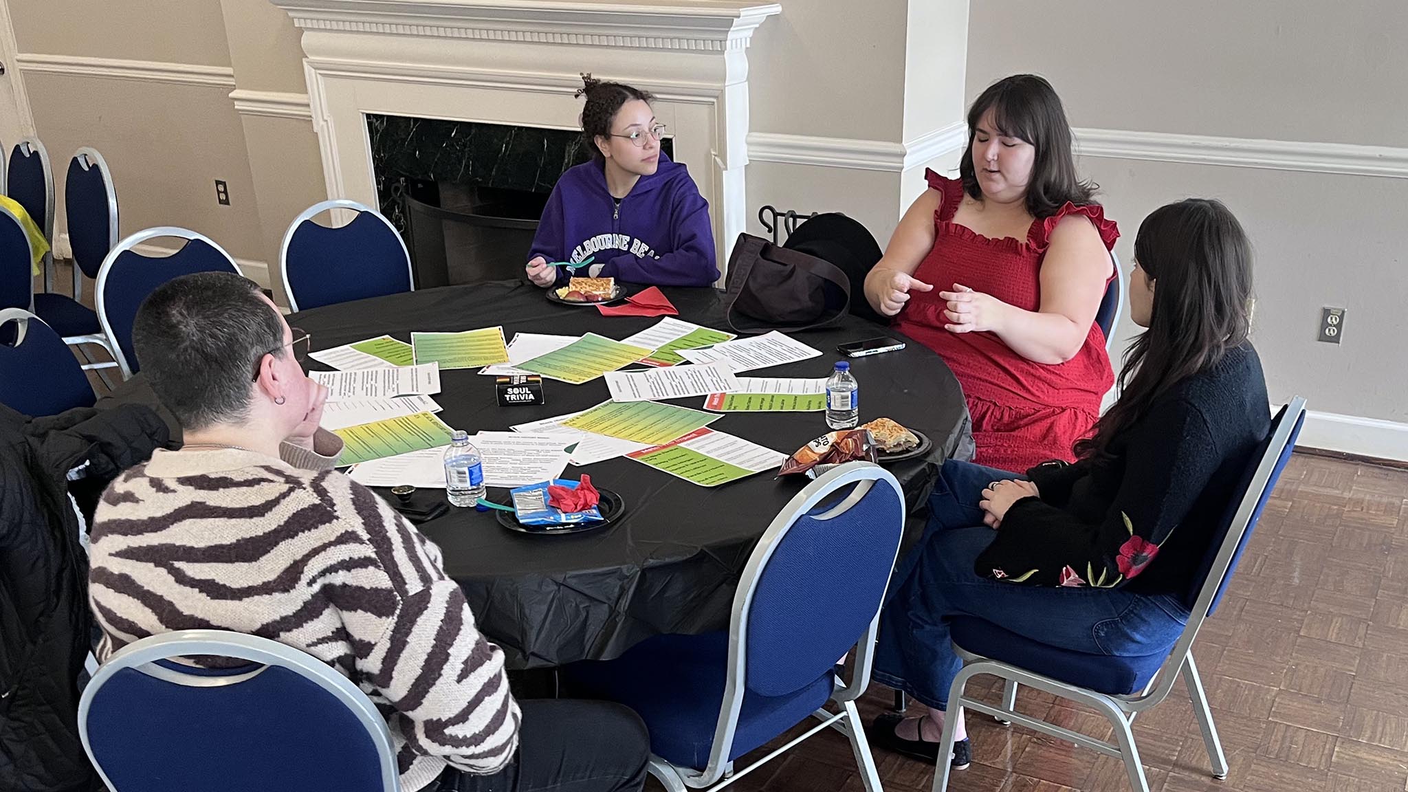 Four people sit around a round table with papers, water bottles, and food in a well-lit room with blue chairs and white walls.