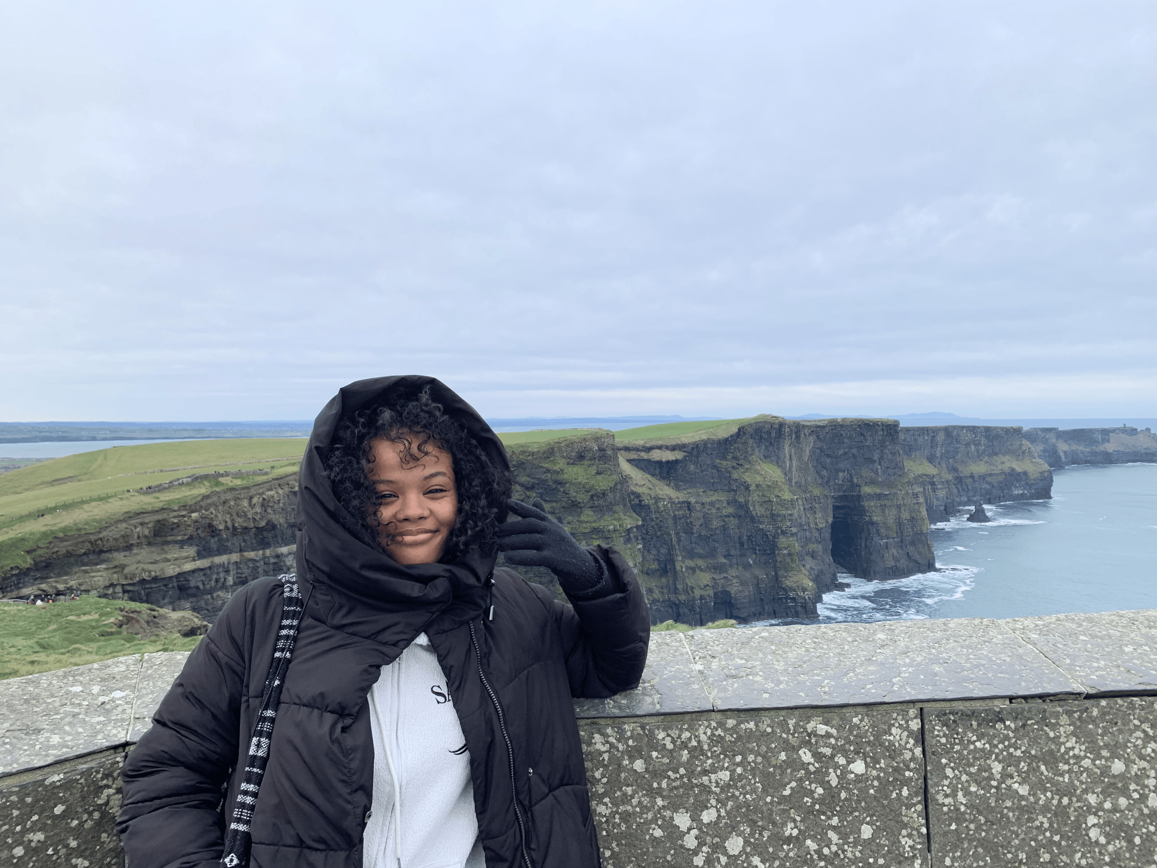 Person in a black and white winter coat stands in front of the Cliffs of Moher, Ireland, with cloudy skies and cliffs visible in the background.