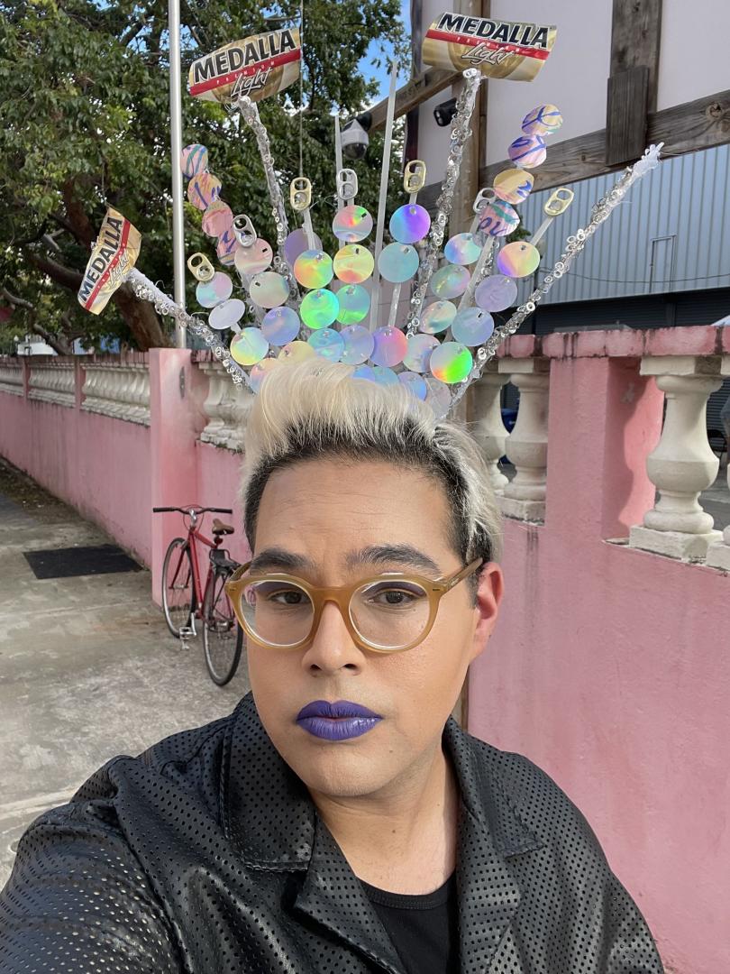 Person with light hair and purple lipstick wears glasses and an elaborate headpiece made of shiny discs, beads, and Medalla Light beer cans, standing outside near a pink wall and a bicycle.