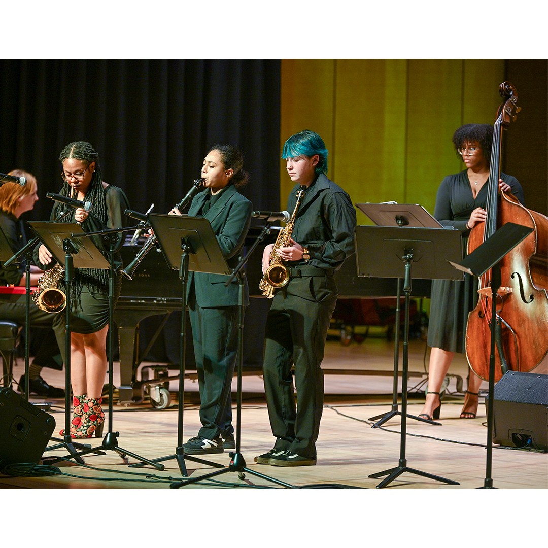 Four musicians perform on stage; three play saxophones while one plays a double bass. All are dressed in black and stand behind music stands.