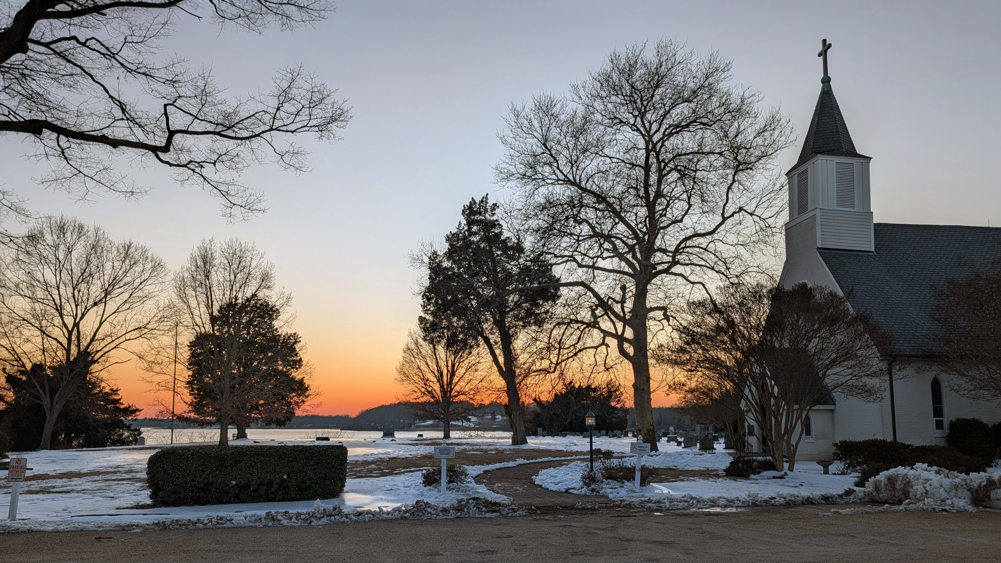 A church with a steeple stands near leafless trees and snow-covered ground at sunset, with a river and distant hills in the background.