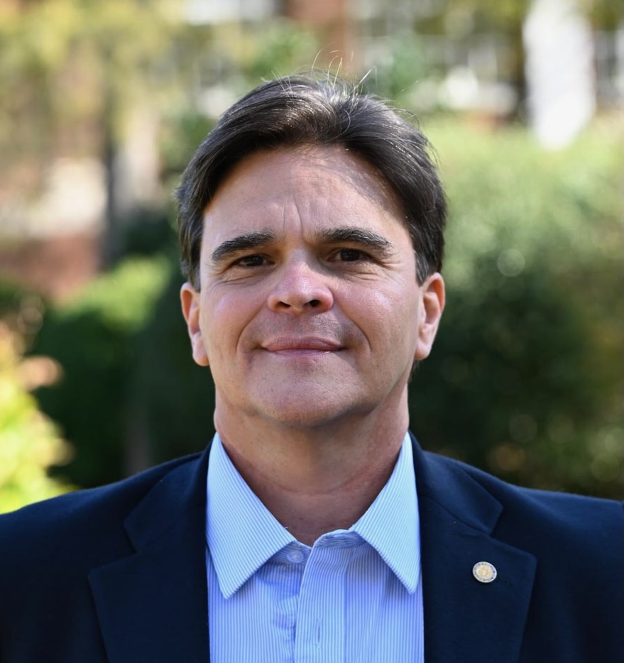 A man in a dark blazer and light blue shirt stands outside, facing the camera, with greenery and blurred buildings in the background.