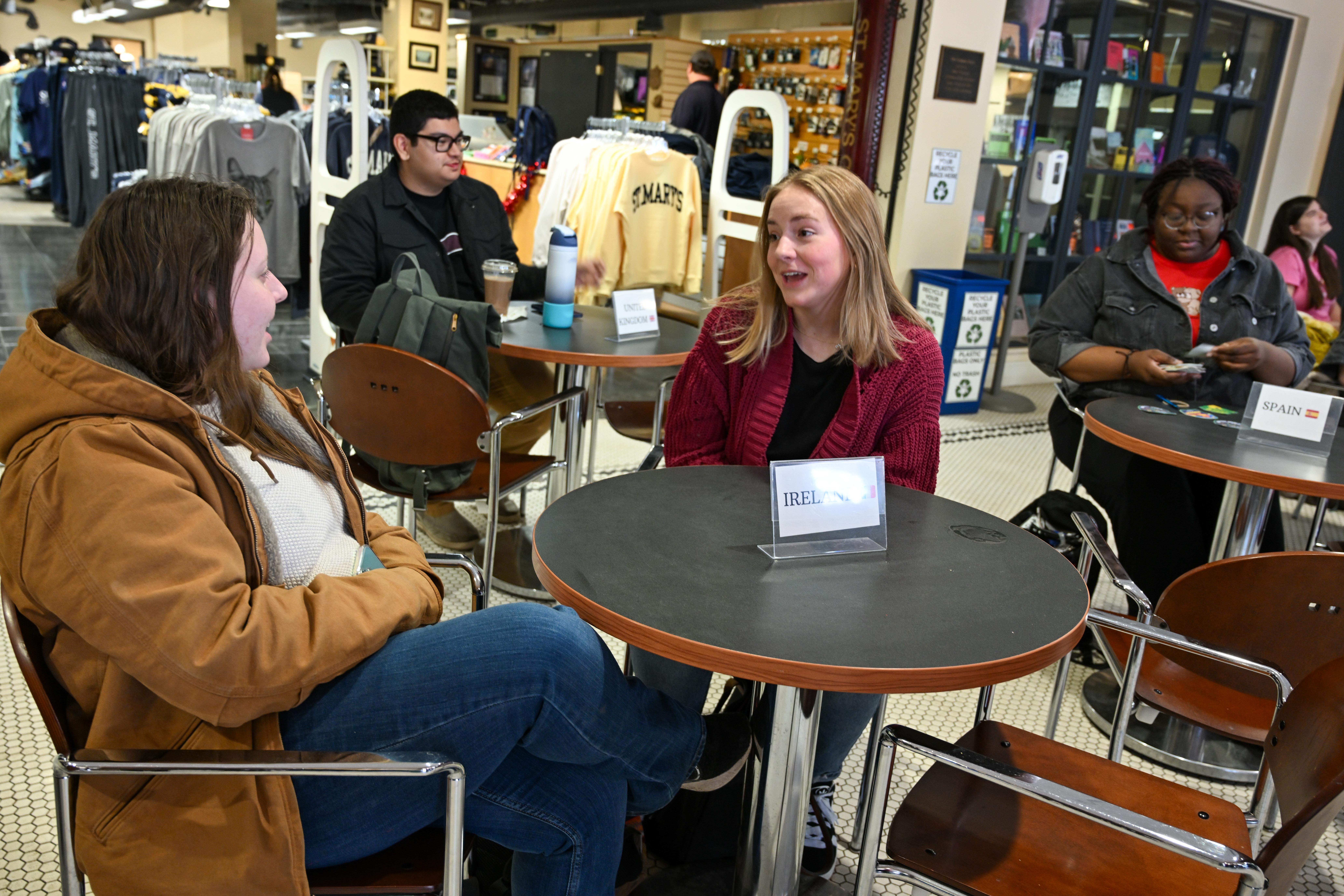 Four students sit at small round tables in a campus store or café. Two women in the foreground talk, while two others use laptops in the background. Shelves of merchandise are visible.