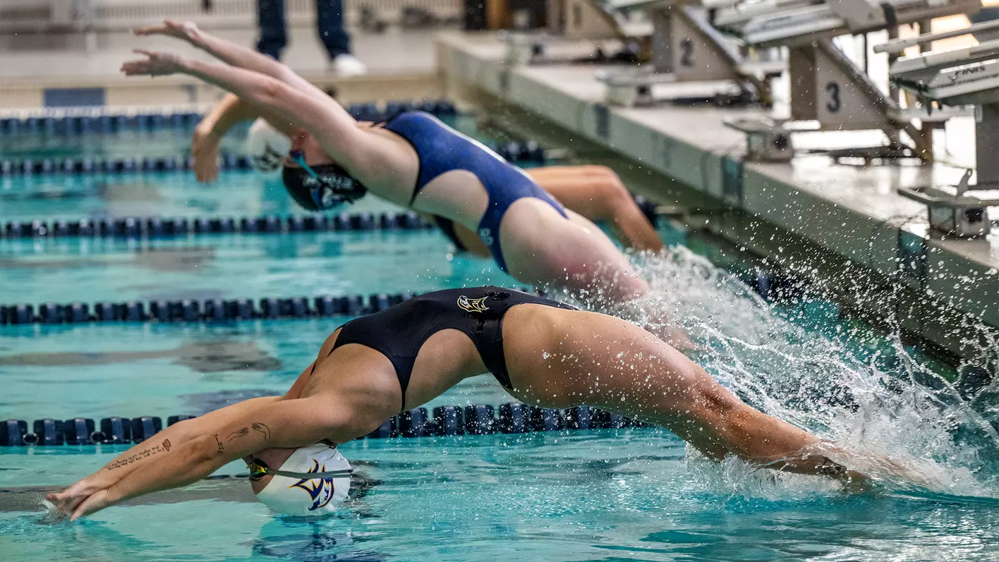 Two competitive swimmers in swimsuits dive backwards into the pool at the start of a backstroke race, with splashes of water visible.