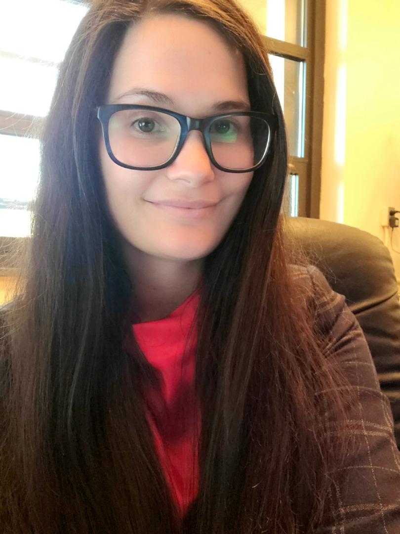 A woman with long brown hair and glasses sits indoors, smiling at the camera. She is wearing a red top and dark blazer.