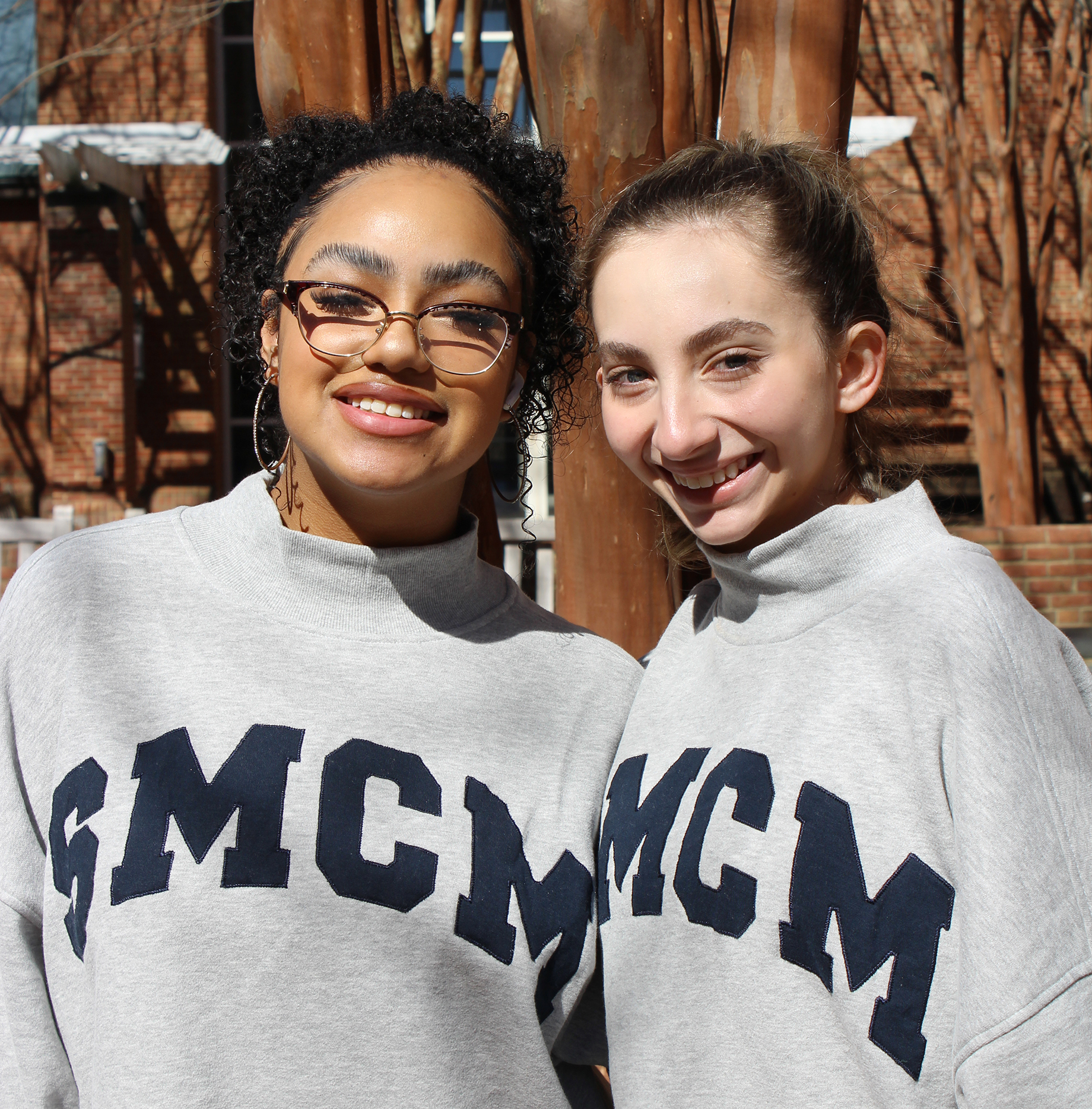 Two young women wearing matching gray sweatshirts with "SMCM" lettering stand outdoors, smiling at the camera.