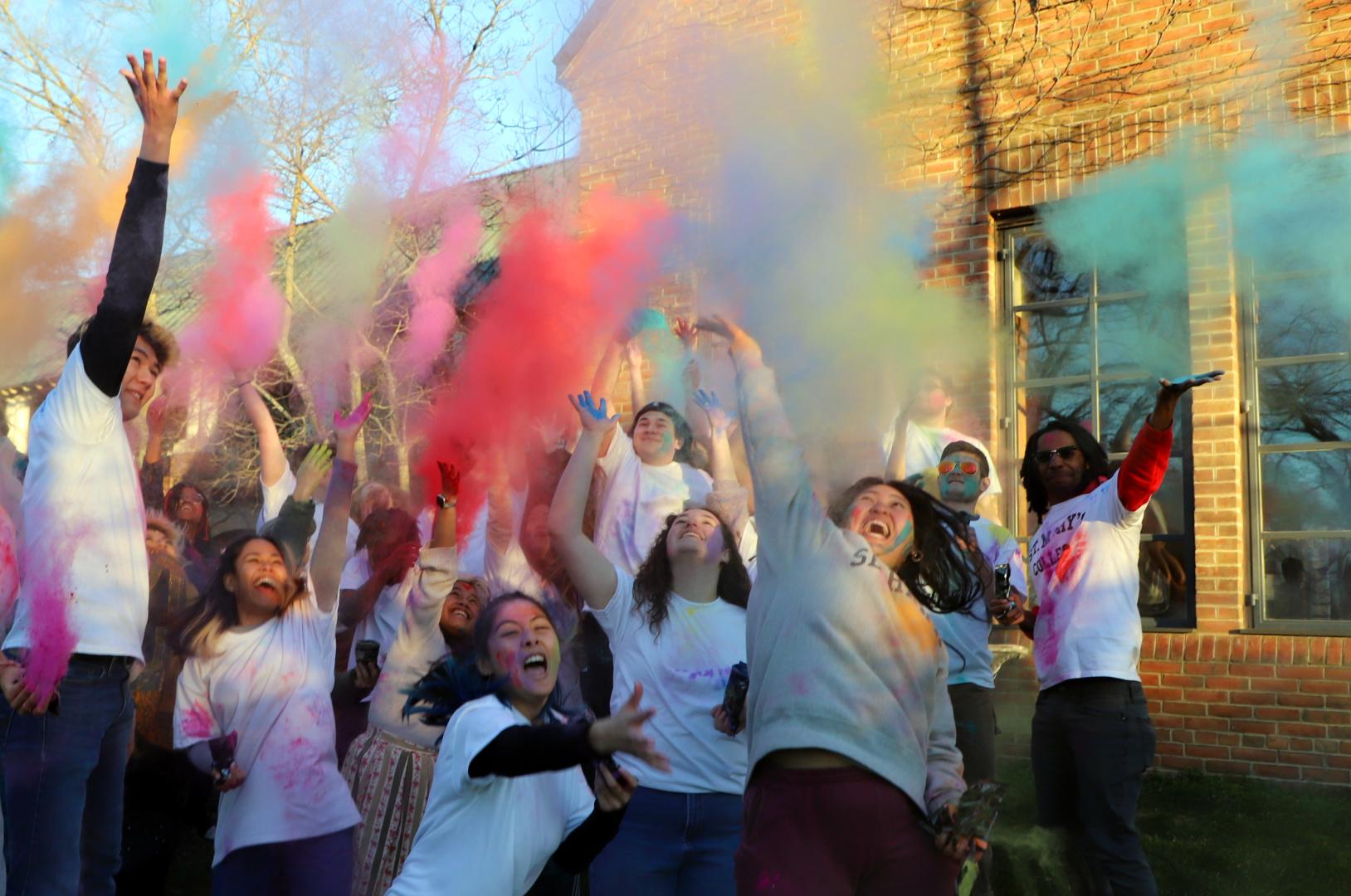 A group of people outdoors throw colorful powder into the air, wearing white shirts and smiling in front of a brick building.